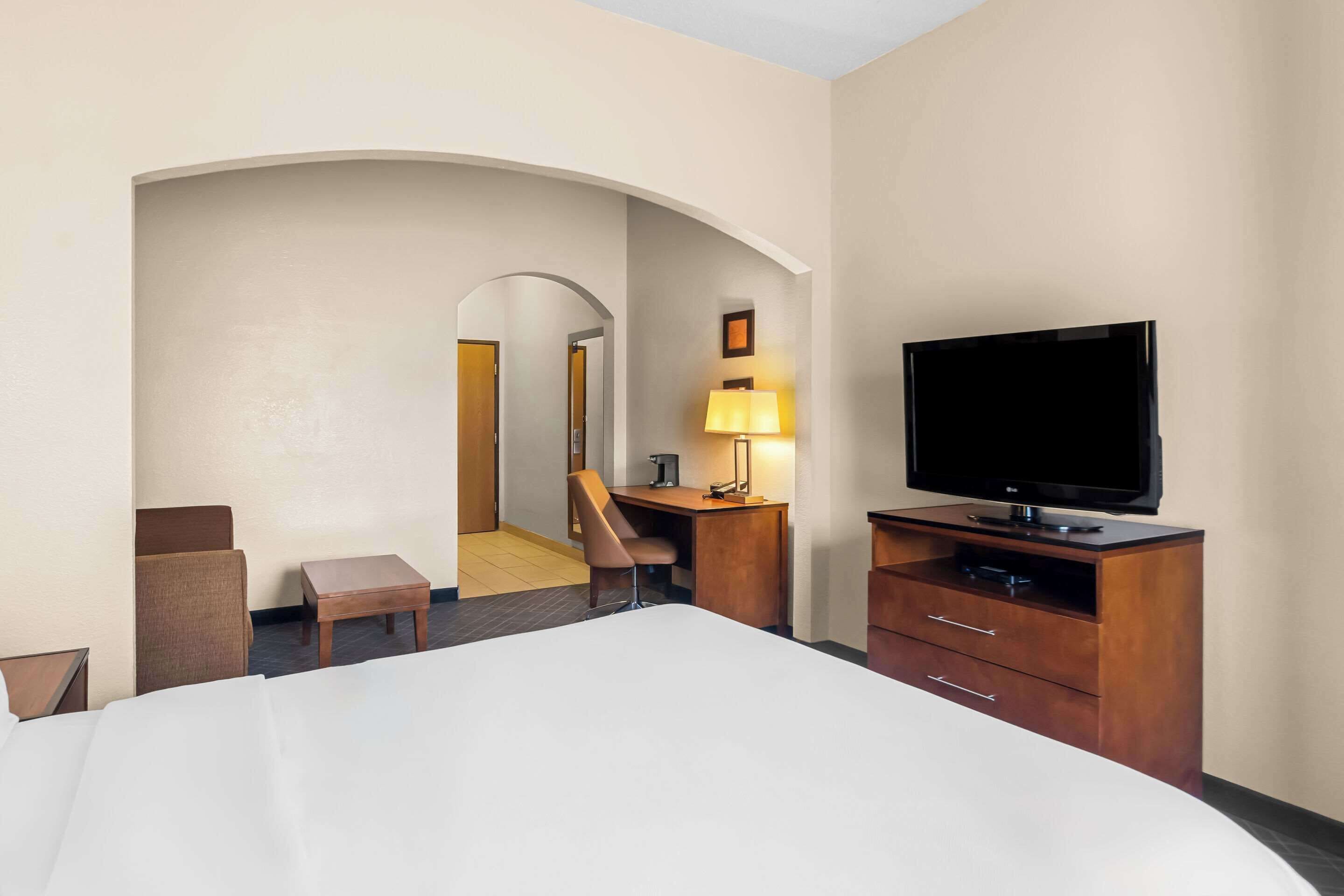 Habitación de hotel con una cama grande, un escritorio con lámpara, televisión y un arco que da a otra habitación.
