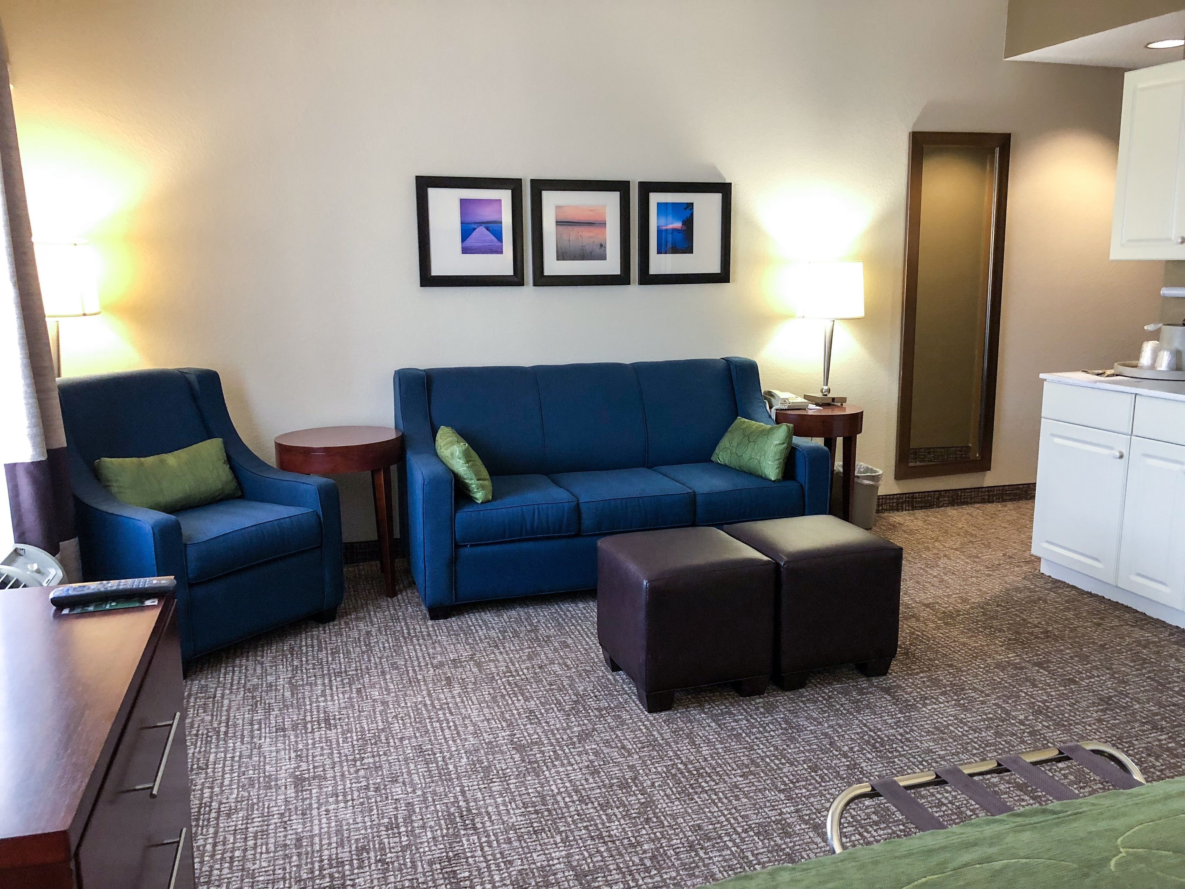 A hotel room with a blue sofa, a matching armchair, a small round table, a lamp, a trash bin, and a kitchenette area.