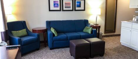 A hotel room with a blue sofa, a matching armchair, a small round table, a lamp, a trash bin, and a kitchenette area.