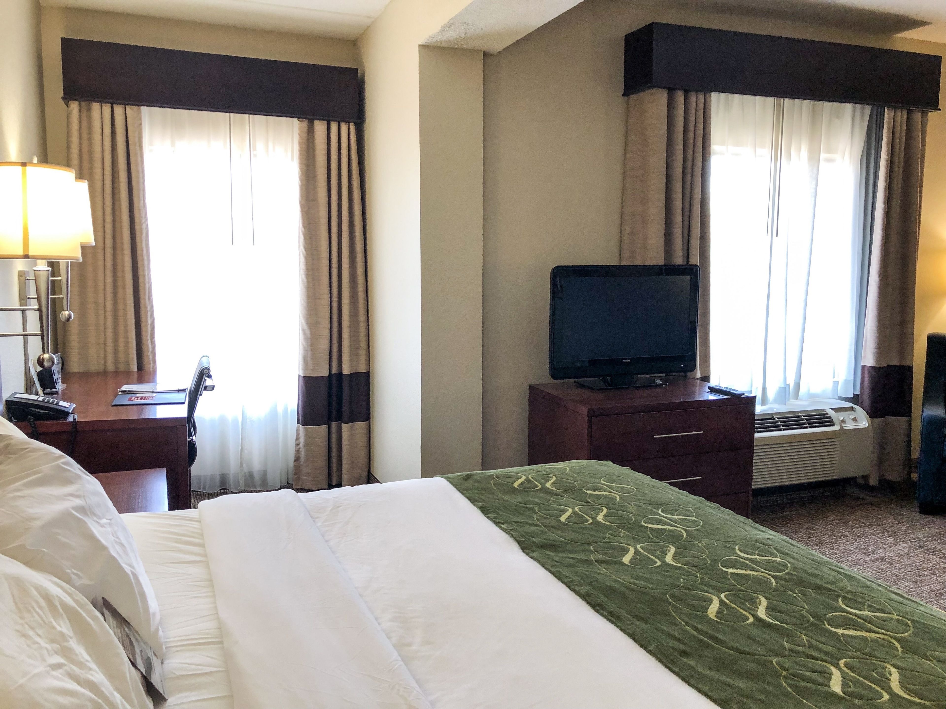 A hotel room with a bed, a desk with a lamp, a television, and an air conditioning unit.