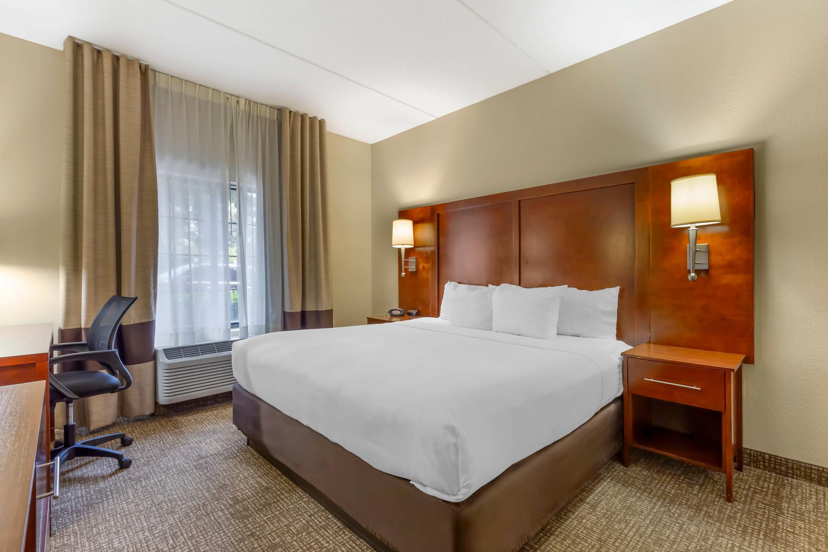 A hotel room with a large bed, a desk with a chair, a nightstand with a lamp, and a window with curtains.