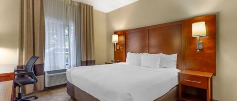 A hotel room with a large bed, a desk with a chair, a nightstand with a lamp, and a window with curtains.