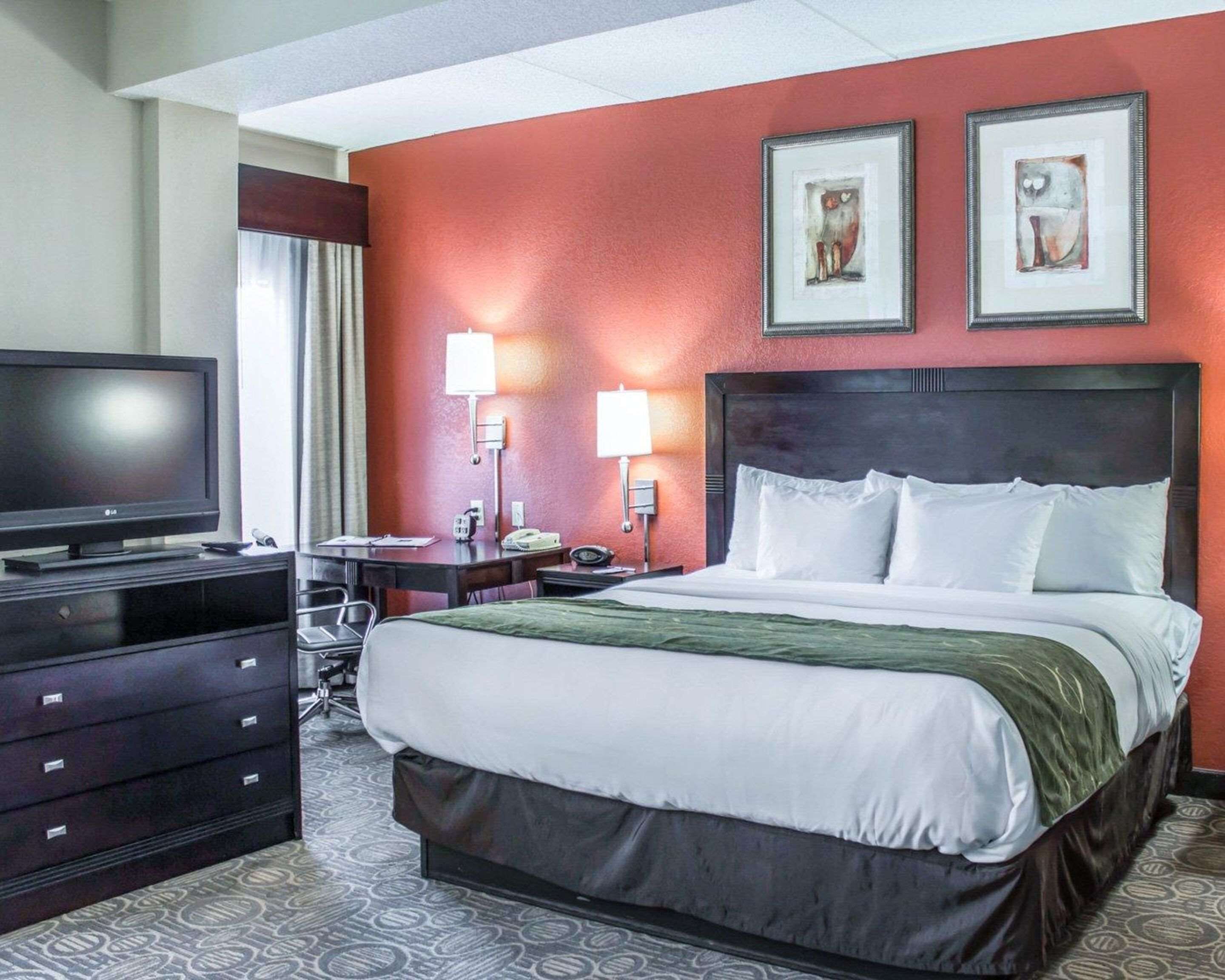 Habitación de hotel con cama, televisión, escritorio con silla, dos lámparas de pared y dos obras de arte enmarcadas.