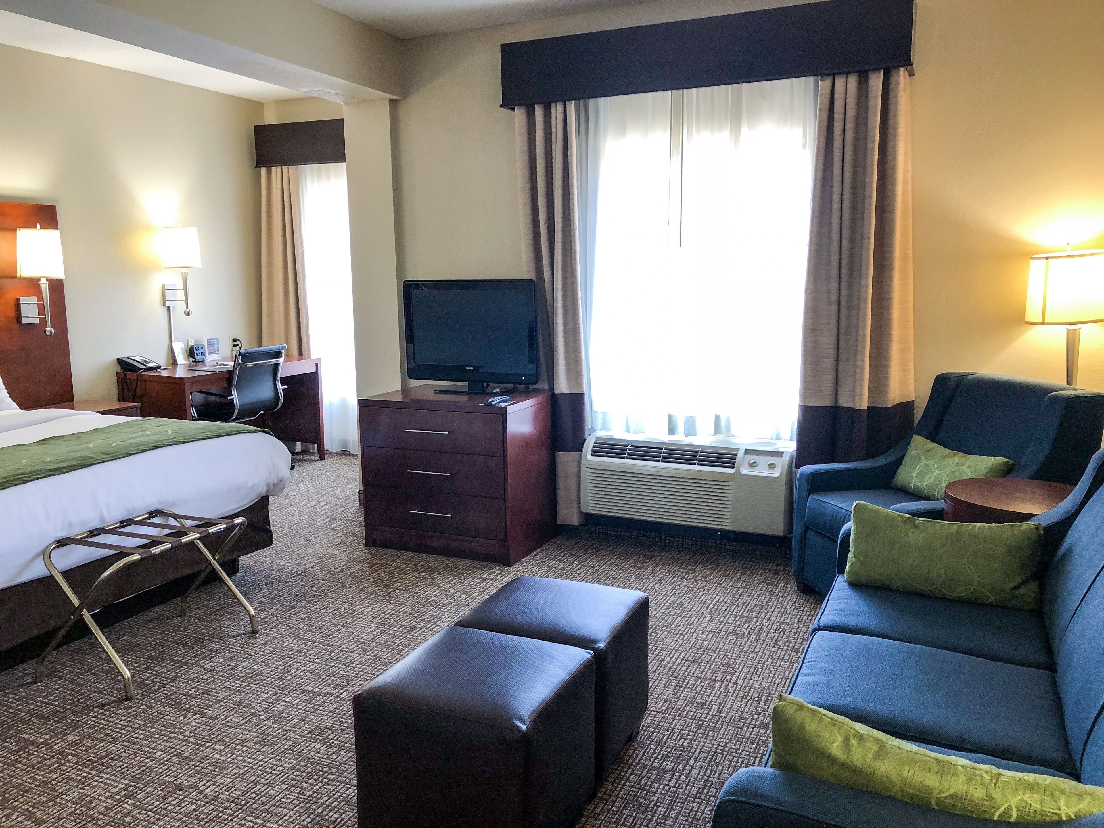 A hotel room with a bed, a desk, a television, a sofa, and an air conditioning unit.