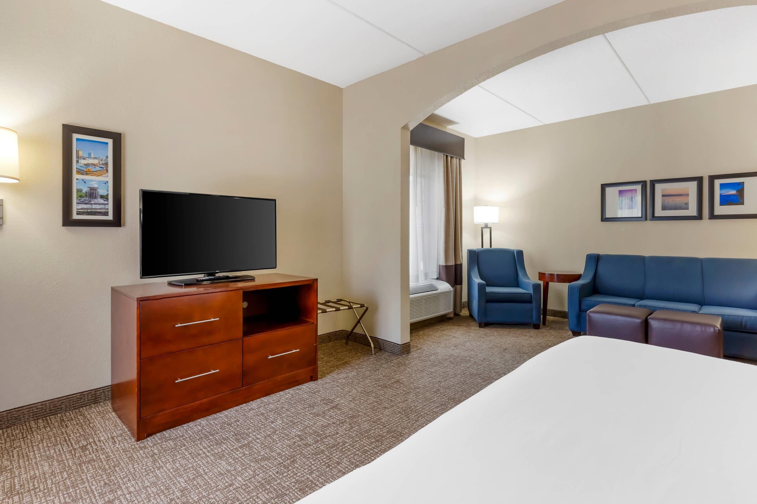 Habitación de hotel con una cama, un televisor sobre un mueble de madera, un sofá azul, una mesita y una ventana con cortinas.
