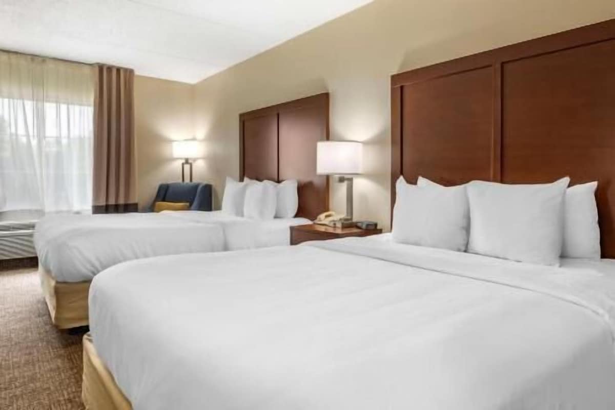 Une chambre d’hôtel avec deux lits, un bureau avec une lampe, une chaise et une fenêtre avec des rideaux.