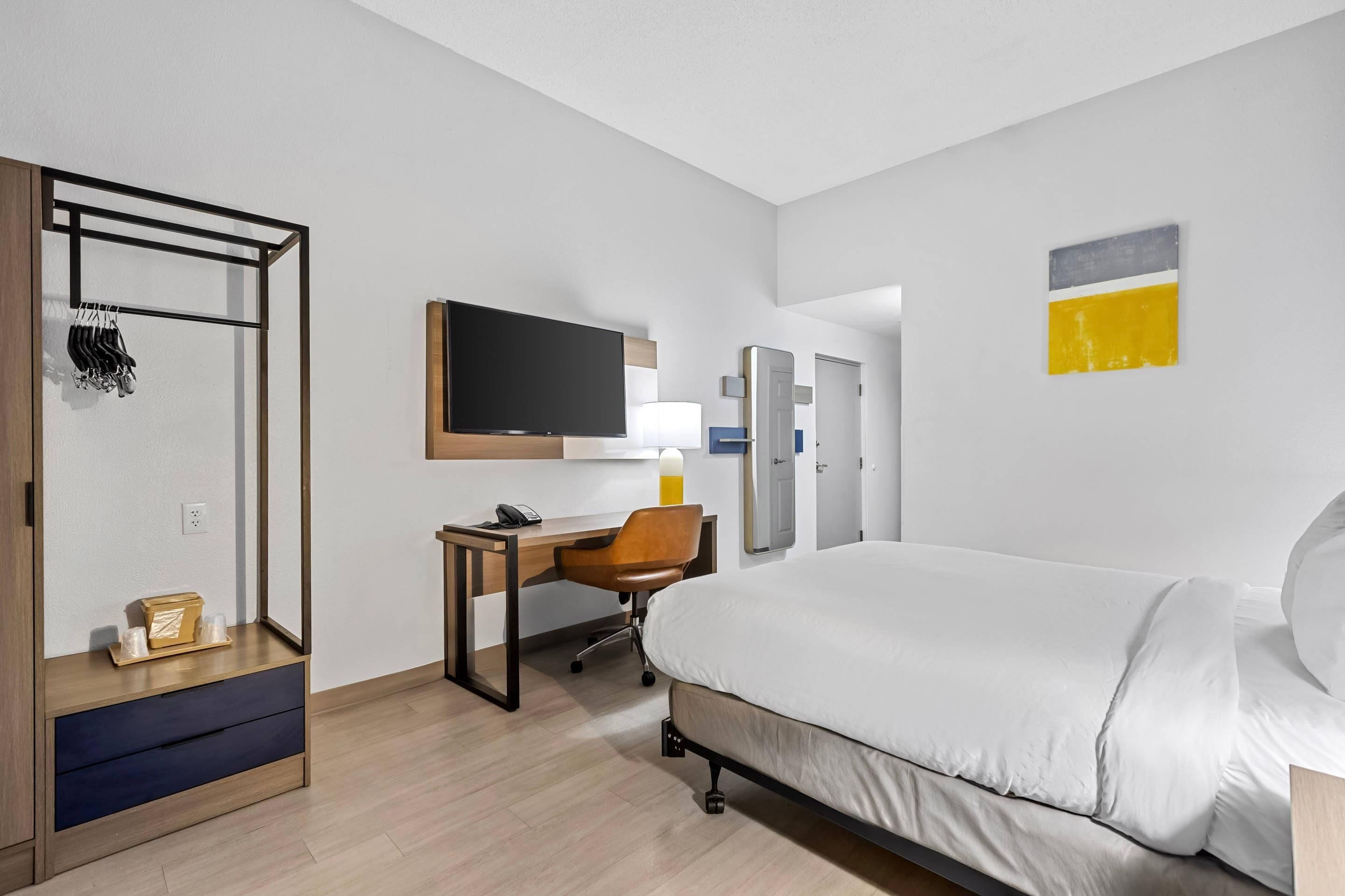 Una habitación de hotel con cama, escritorio, silla, televisor, armario y espejo.