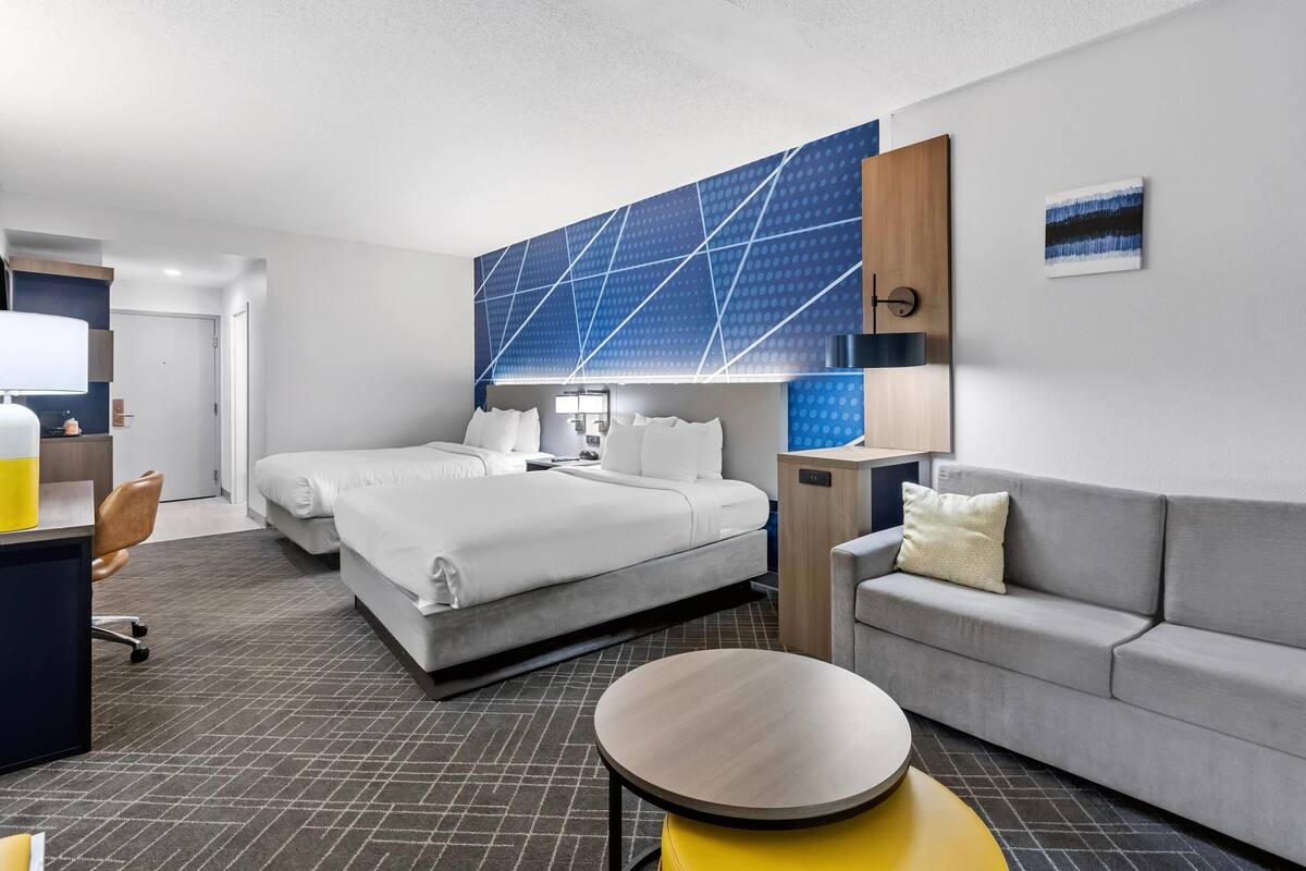 A hotel room with two beds, a sofa, a desk with a chair, and a large wall art piece.