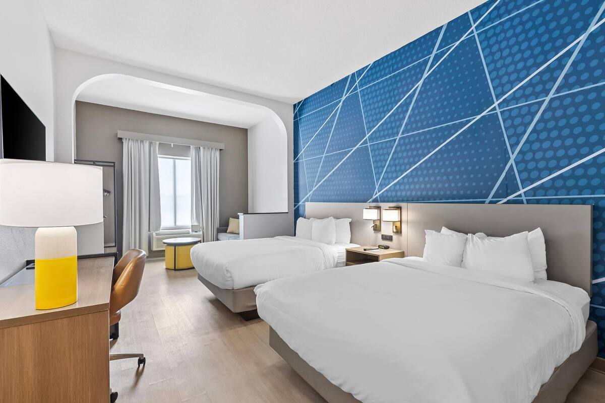 A hotel room with two beds, a desk, a chair, a lamp, and a large window with curtains.