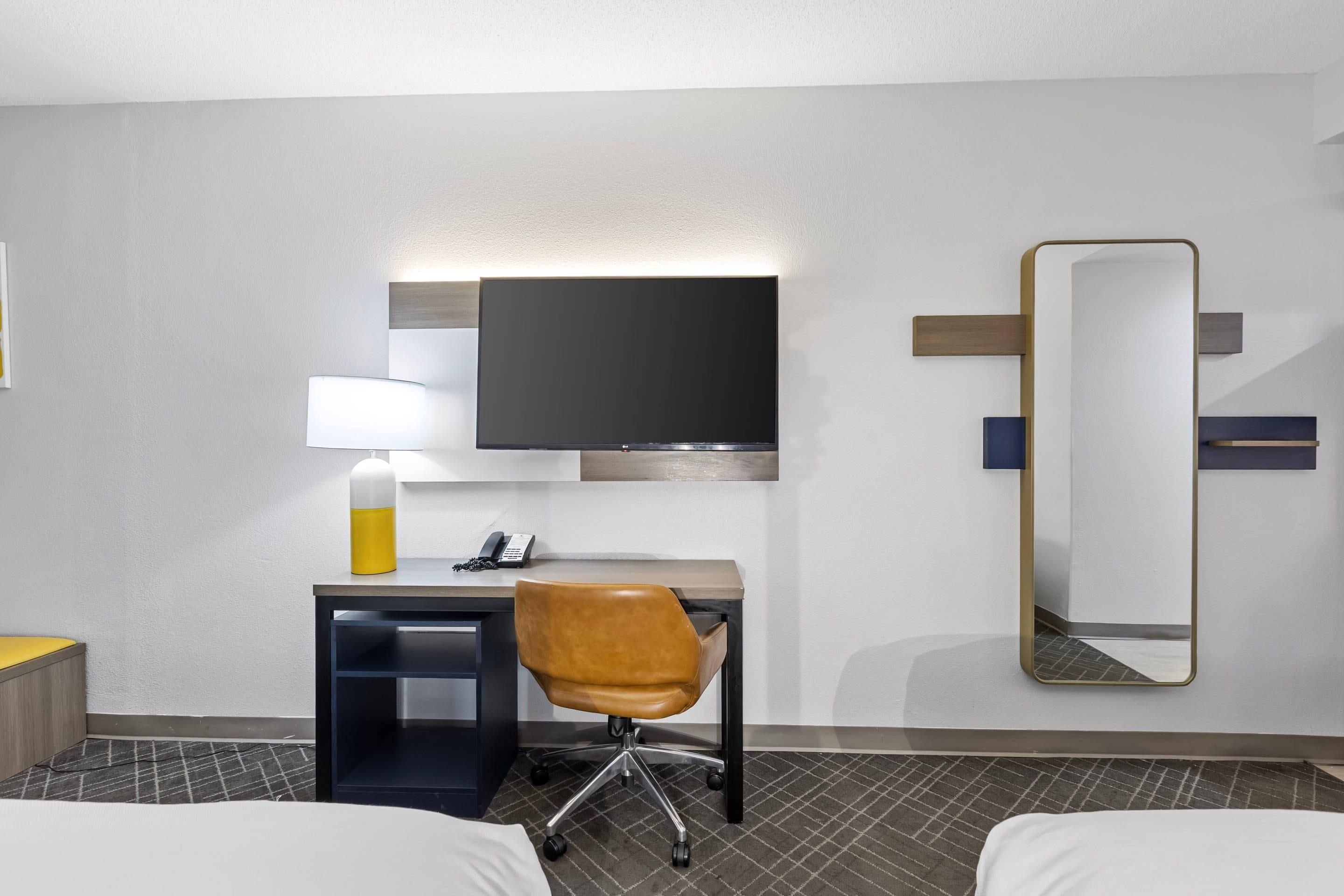A hotel room with a bed, a desk with a chair, a flat-screen TV, a bedside lamp, a mirror, and a telephone.