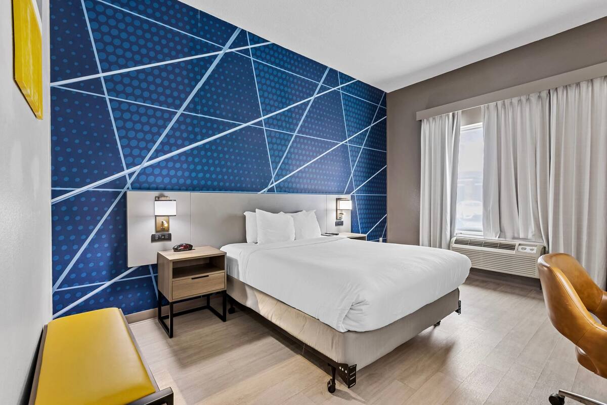 A hotel room with a bed, bedside tables, a chair, a desk, a window with curtains, and a wall with a geometric pattern.