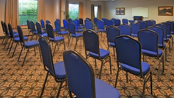 Un salón de conferencias con filas de sillas azules, alfombra estampada y grandes ventanales con cortinas.