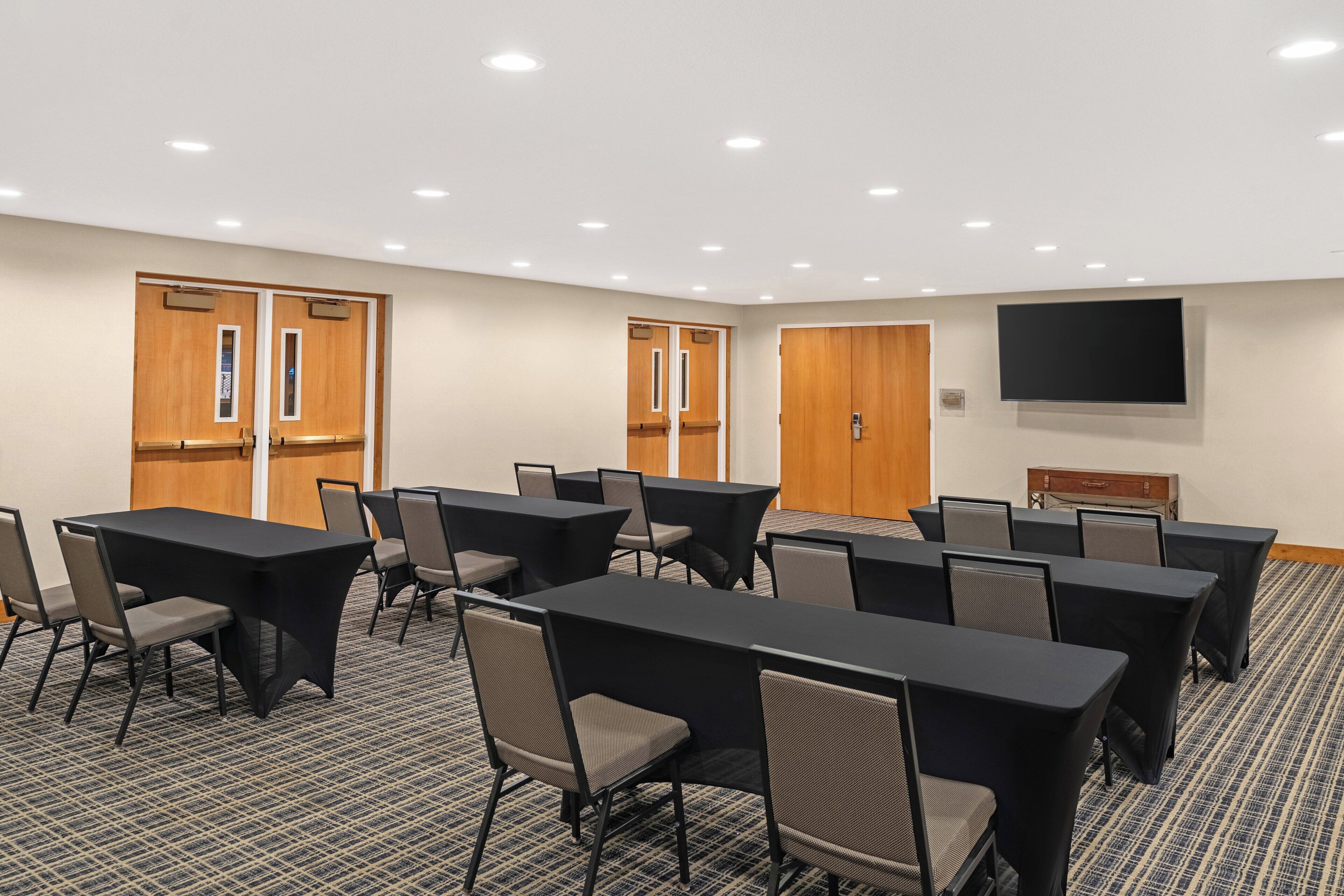 A conference room with multiple tables and chairs, wooden doors, a flat-screen TV, and a carpeted floor.