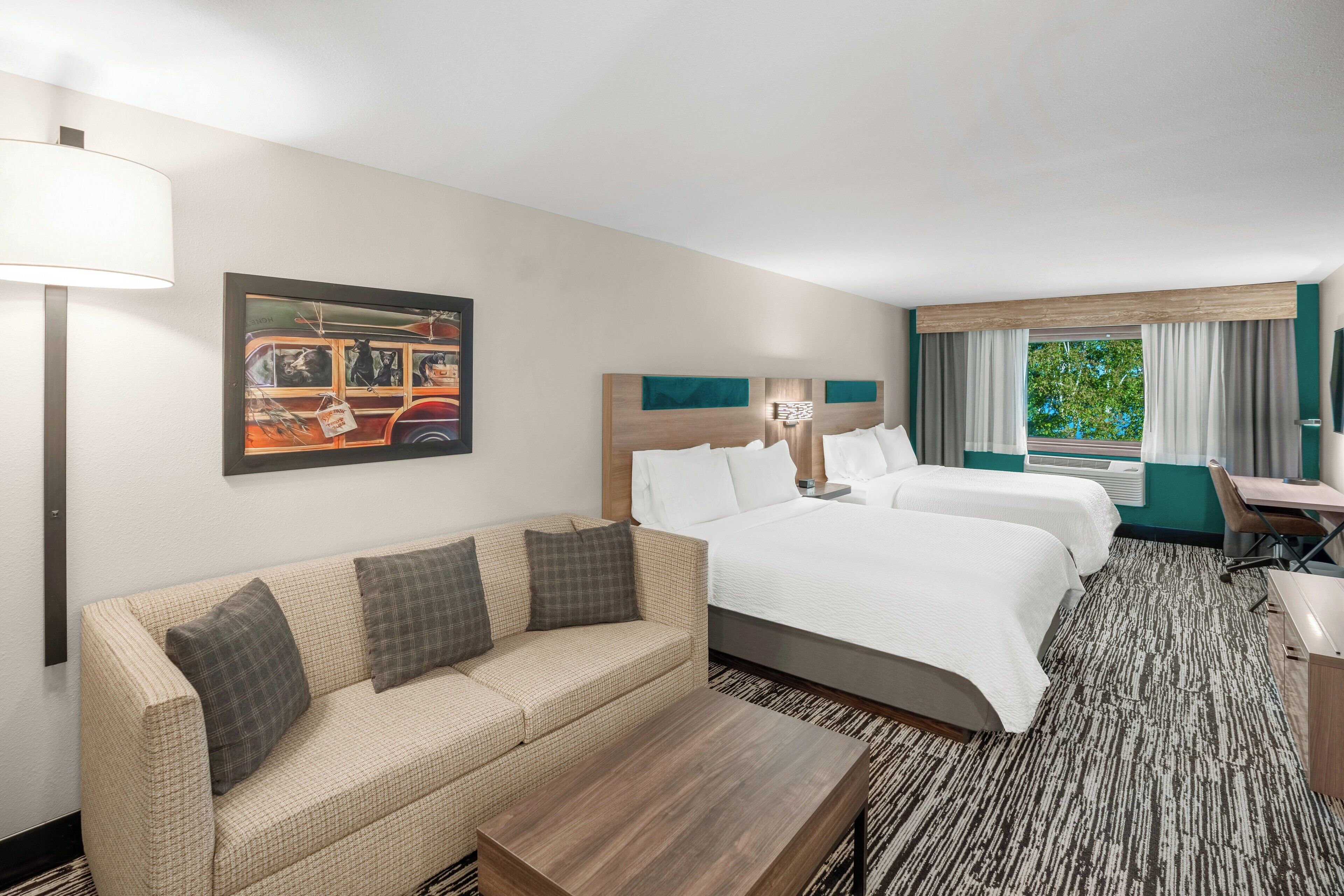 A hotel room with a sofa, two beds, a desk, a lamp, and a painting on the wall.