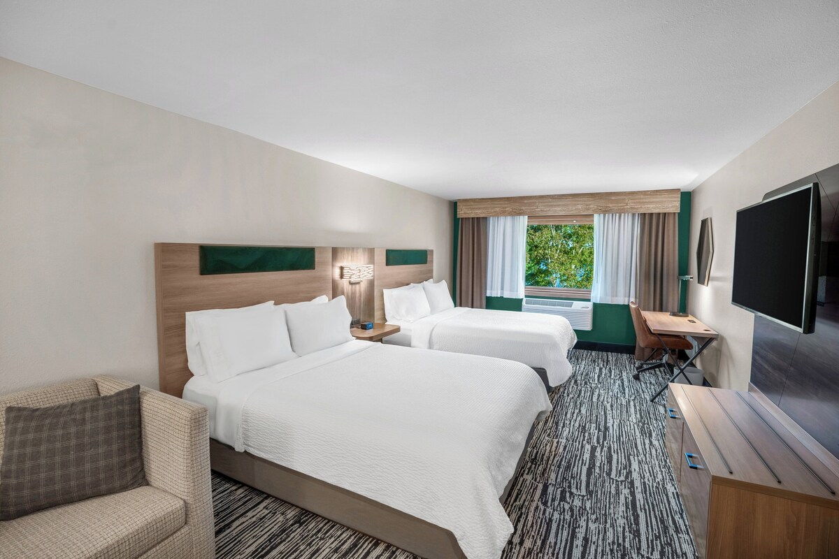 A hotel room with two beds, a desk, a TV, and a view of greenery outside.