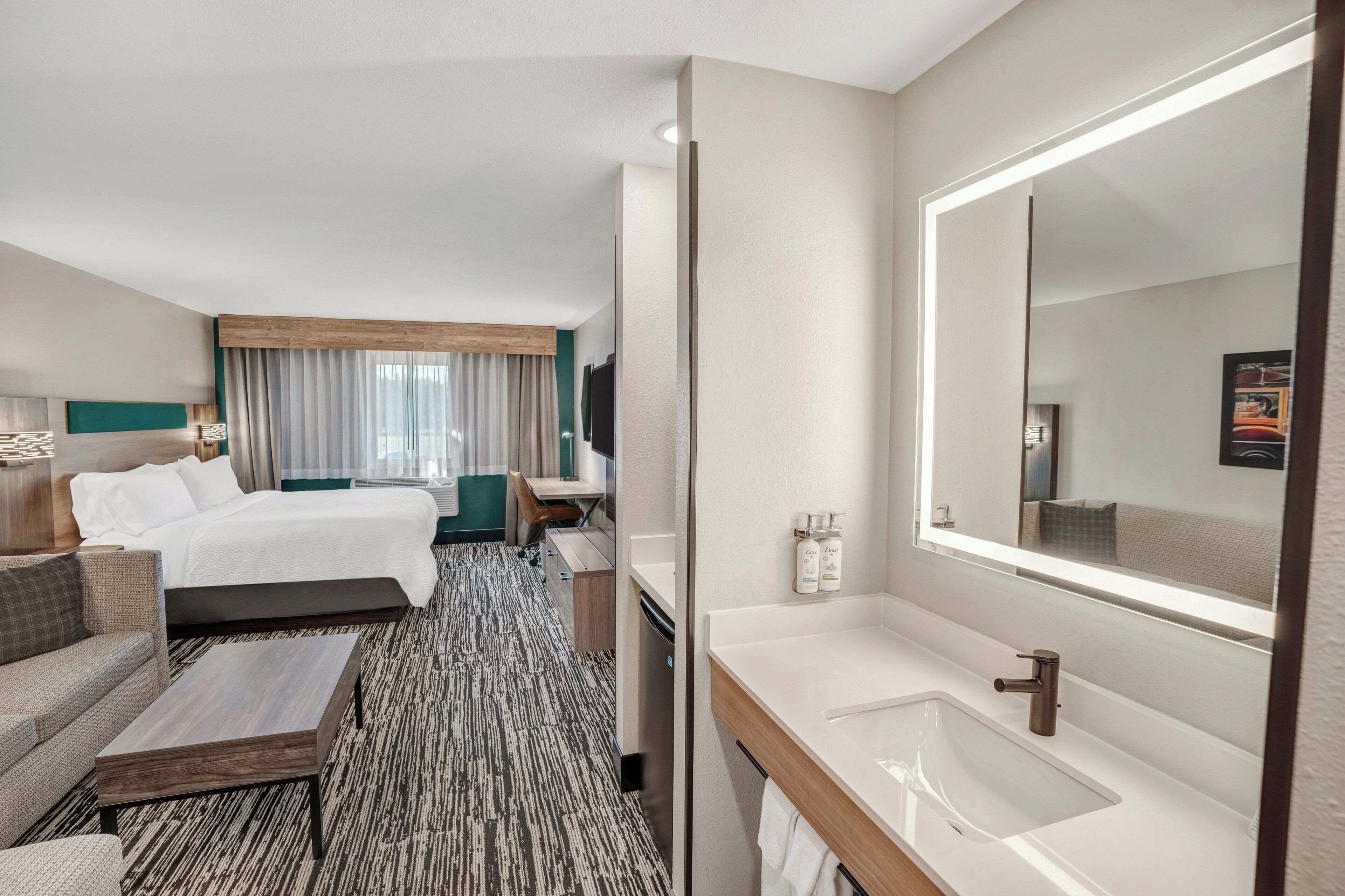 Habitación de hotel con cama, sofá, mesa de centro, baño con lavamanos y espejo, y un ventanal con cortinas.