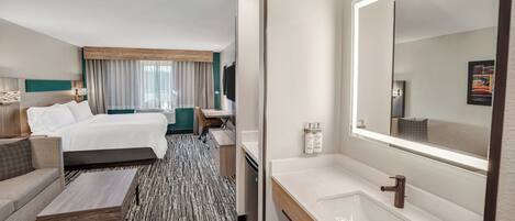 Habitación de hotel con cama, sofá, mesa de centro, baño con lavamanos y espejo, y un ventanal con cortinas.