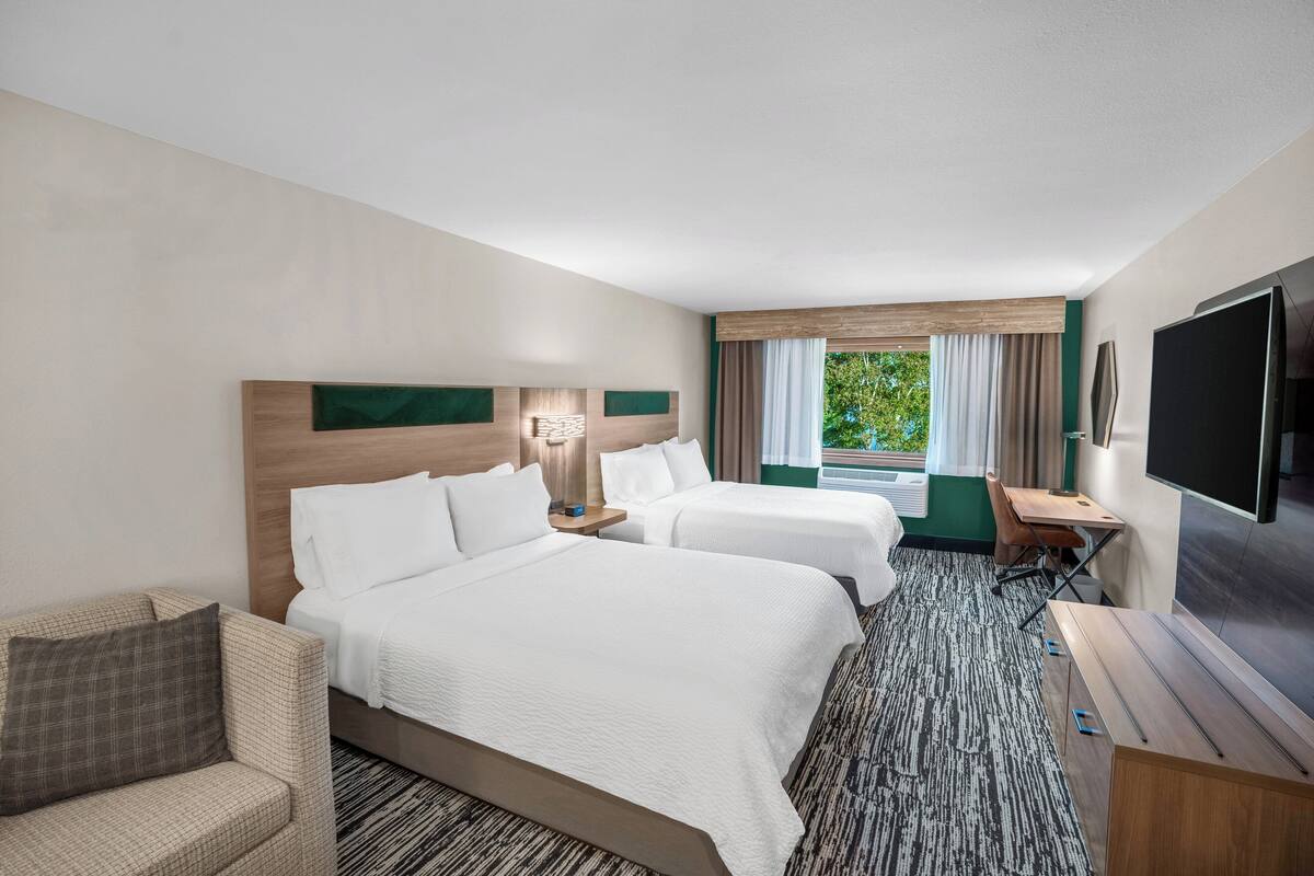 A hotel room with two beds, a desk, a TV, and a view of greenery outside.