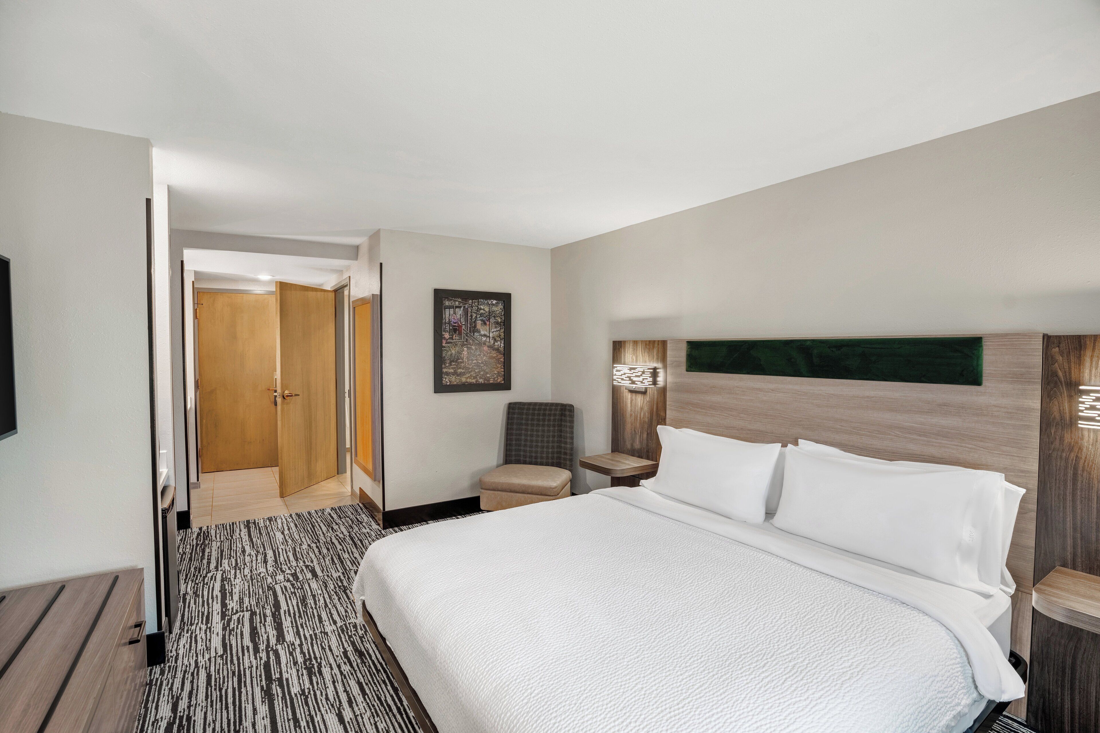 A hotel room with a large bed, a wooden headboard, a small table, a chair, and a door leading to a bathroom.