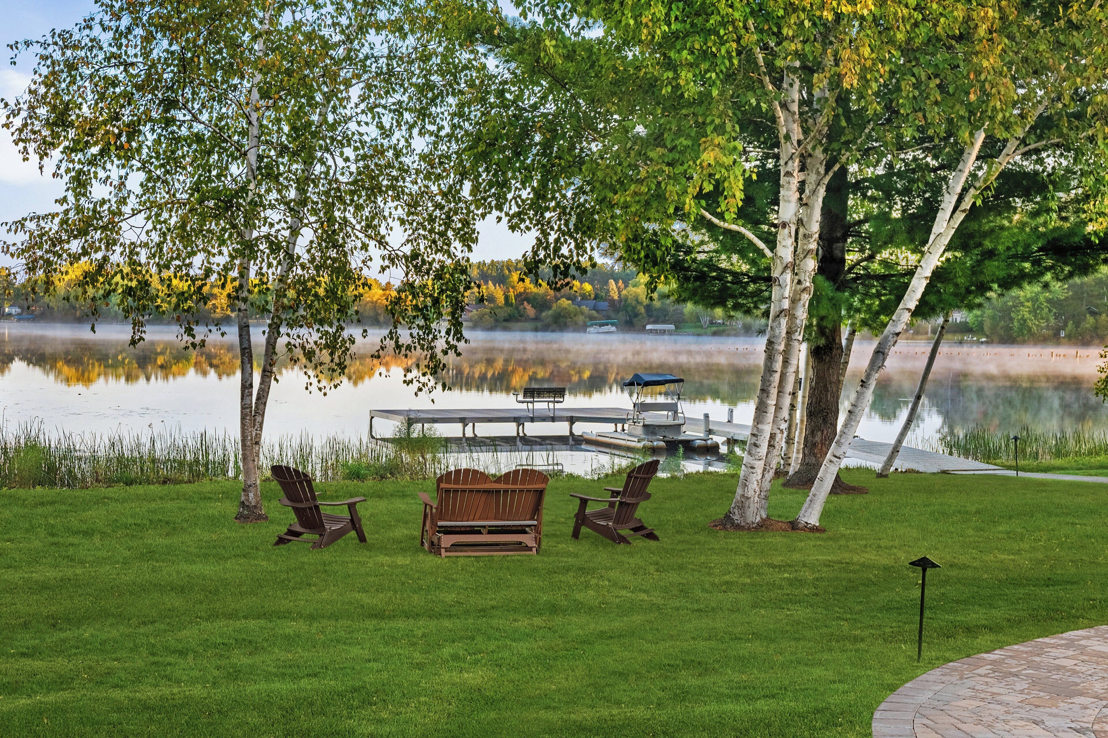 Una terraza junto al lago con sillas Adirondack, un banco de madera y un muelle que se adentra en el agua.