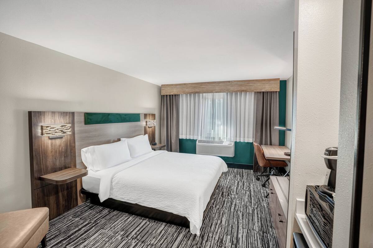 A hotel room with a large bed, a desk, a chair, a TV, and a window with curtains.