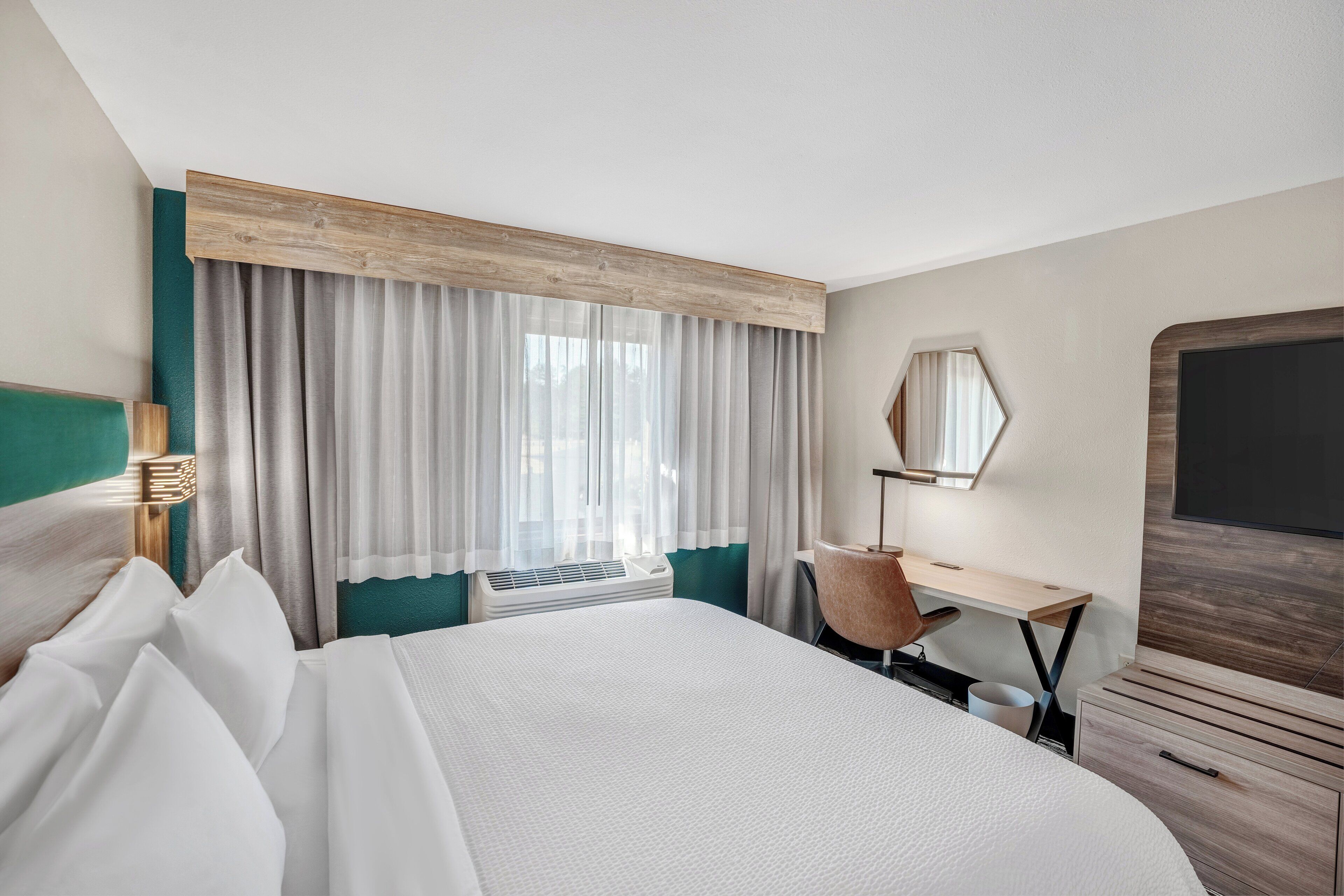 A hotel room with a large bed, a desk, a chair, a TV, and a window with curtains.