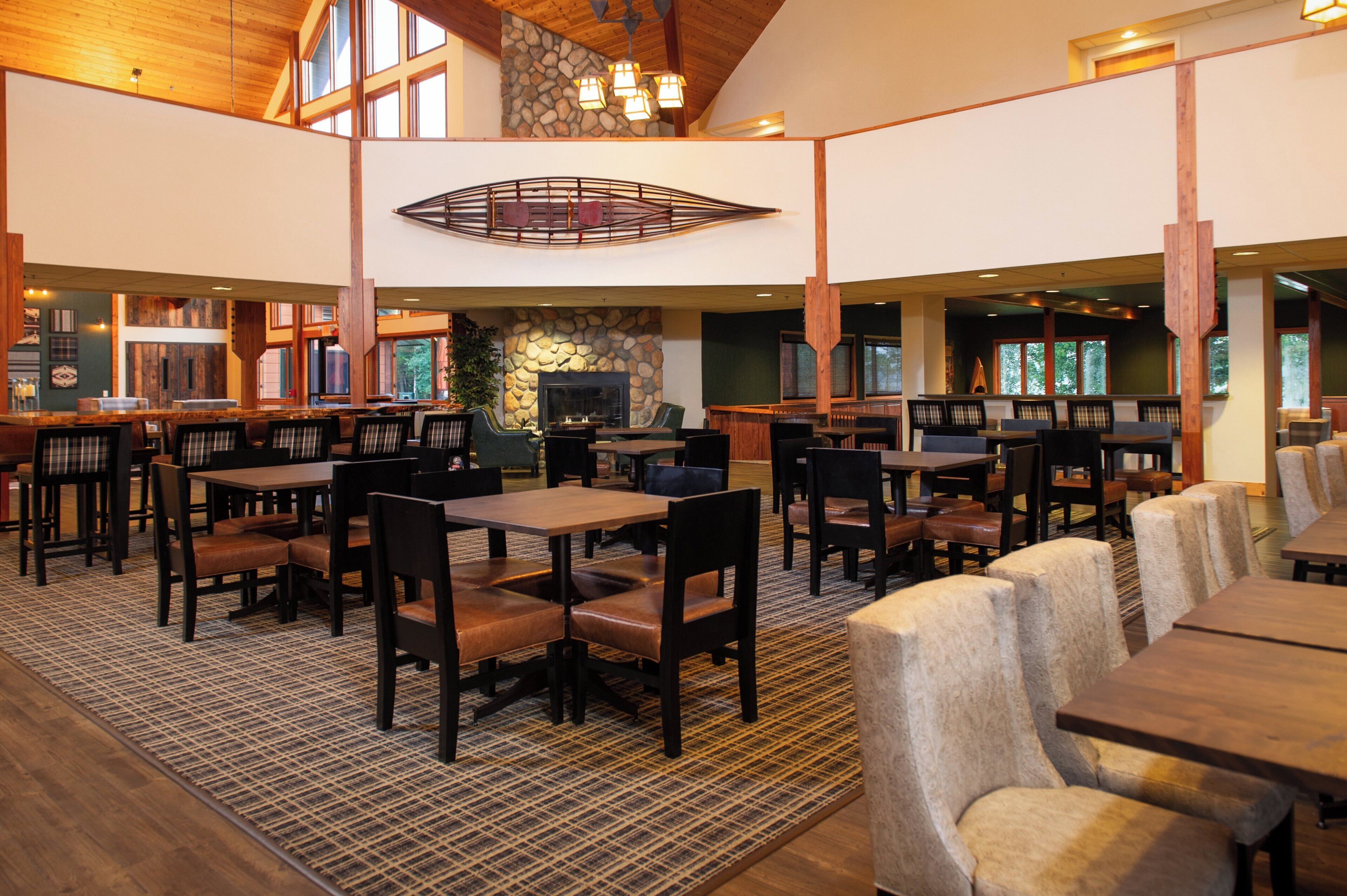A spacious indoor dining area with multiple tables and chairs, a stone fireplace, and large windows allowing natural light.