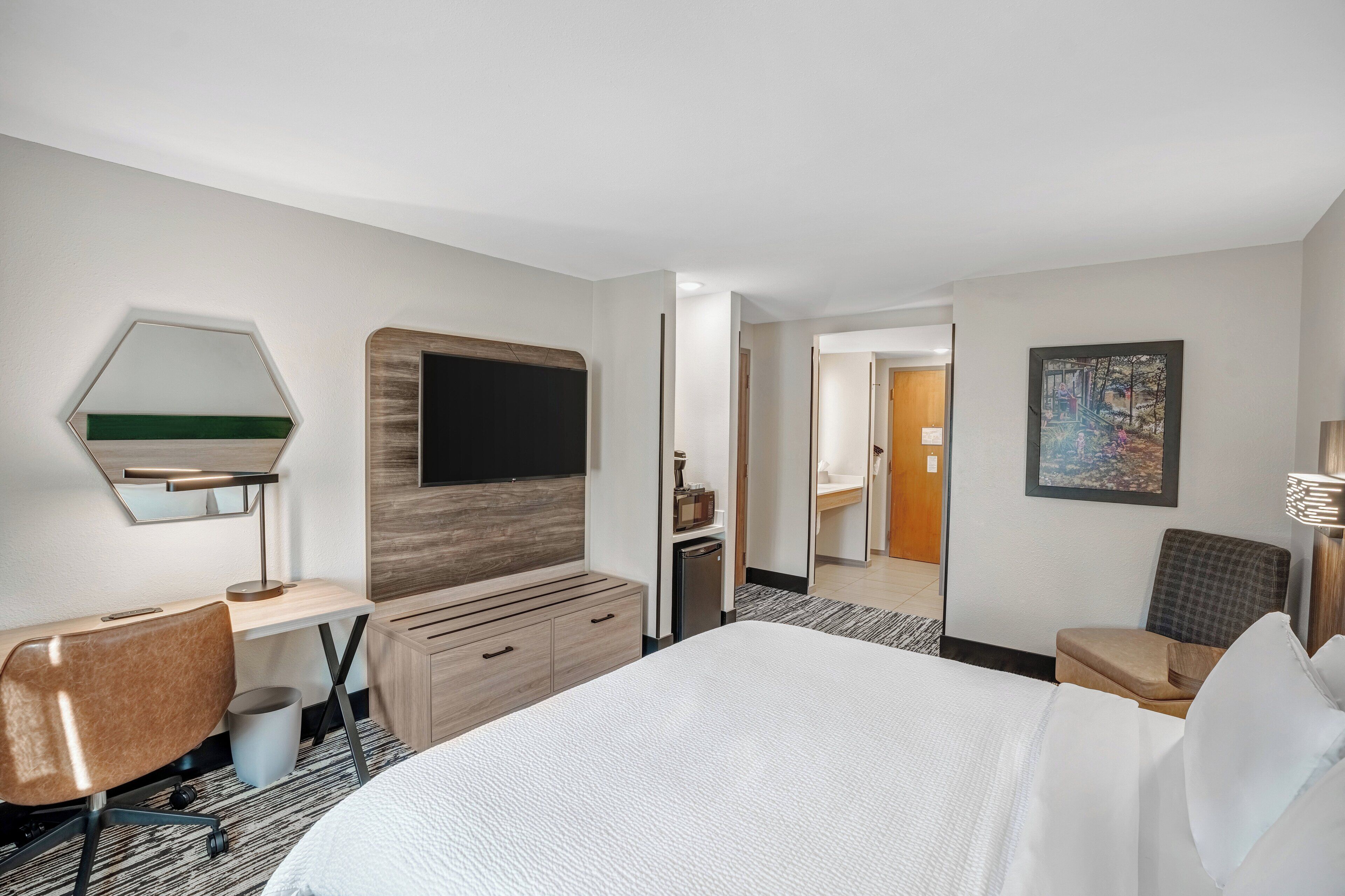 Une chambre d’hôtel avec un grand lit, un bureau avec une chaise, une télévision, un miroir, une commode et un tableau au mur.