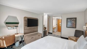 Une chambre d’hôtel avec un grand lit, un bureau avec une chaise, une télévision, un miroir, une commode et un tableau au mur.