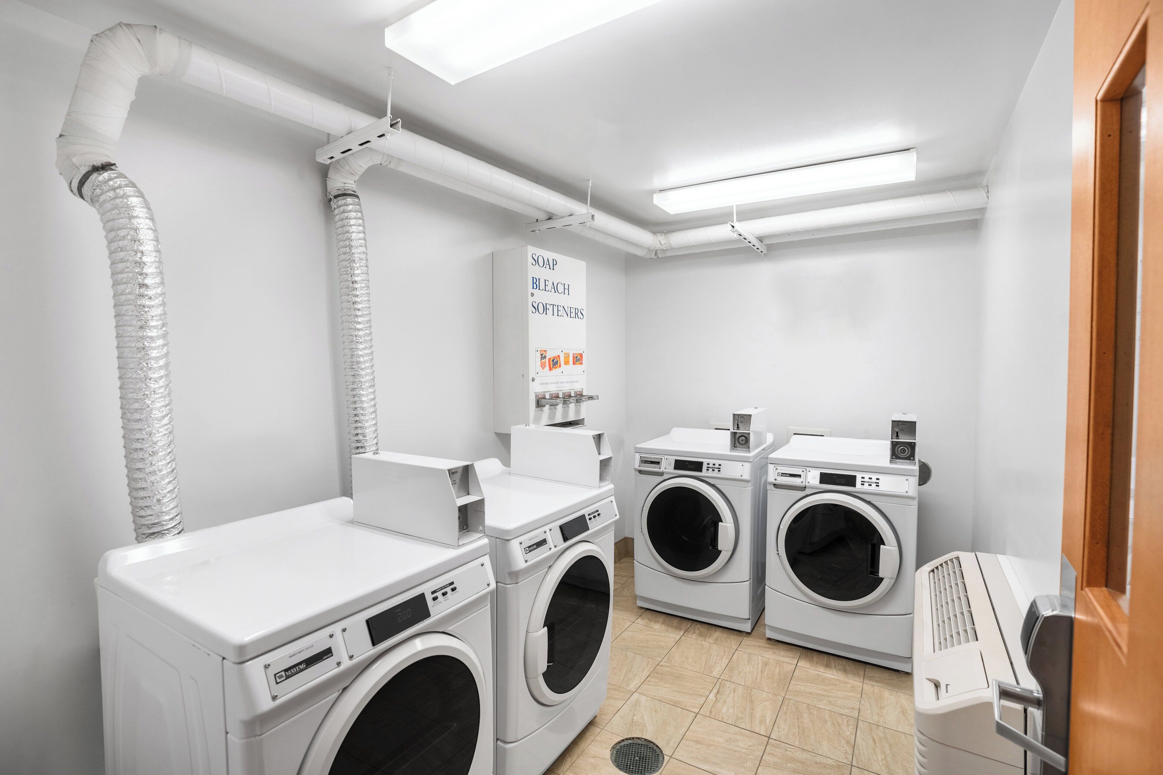A laundry room with three washing machines, a sign for soap and bleach softeners, and ventilation ducts.