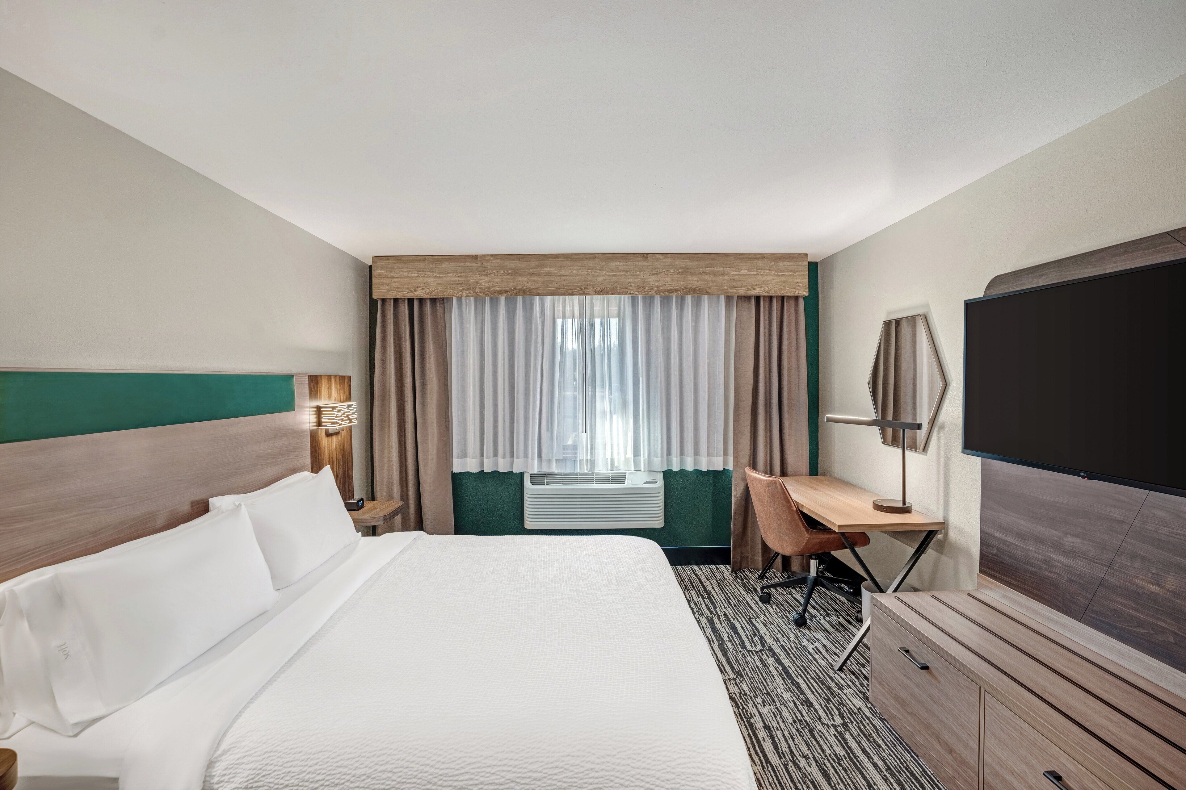 A hotel room with a large bed, a desk, a chair, a flat-screen TV, and a window with curtains.
