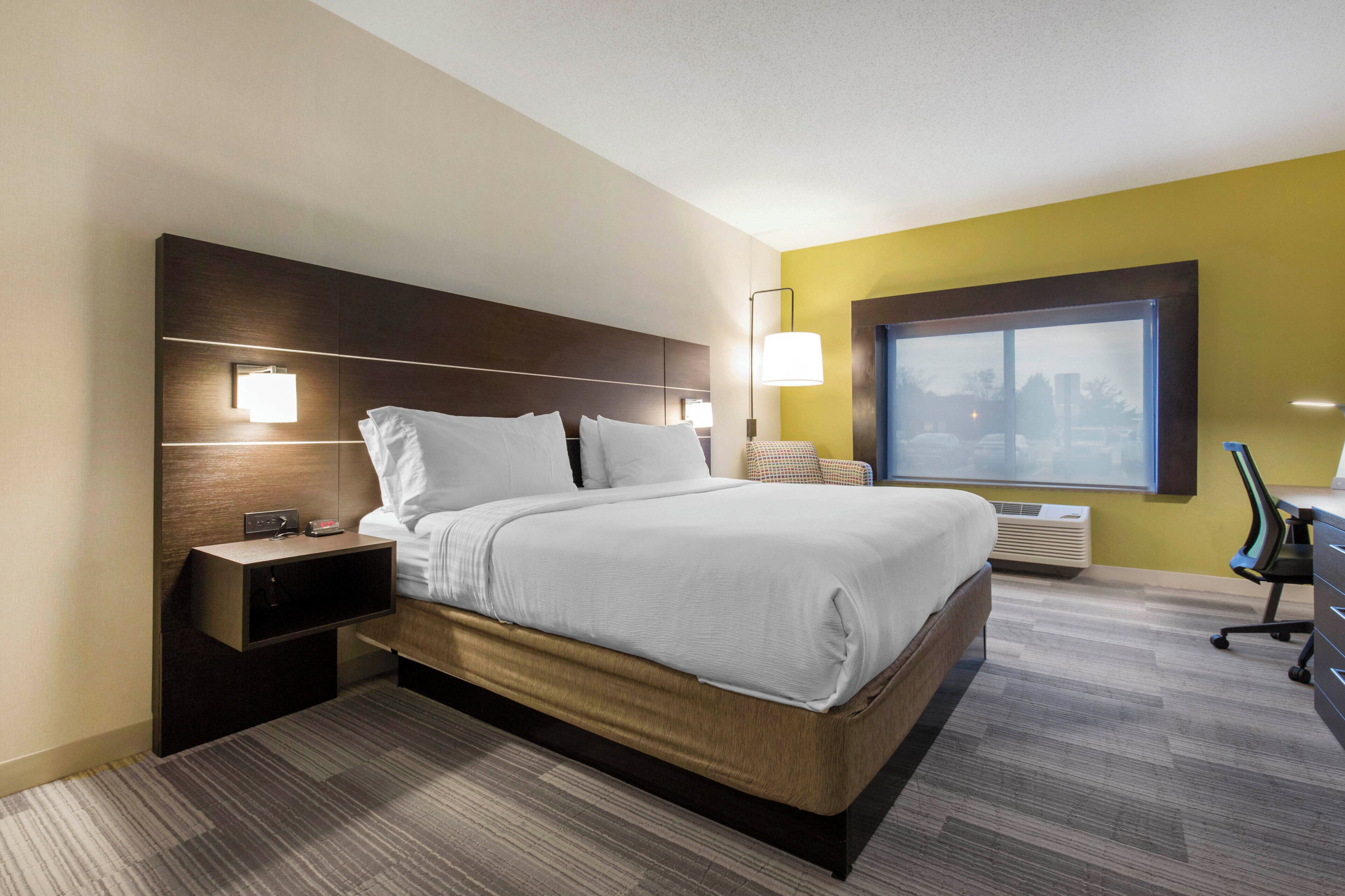 Habitación de hotel con cama, mesita de noche, lámpara y ventana con vista a la ciudad.