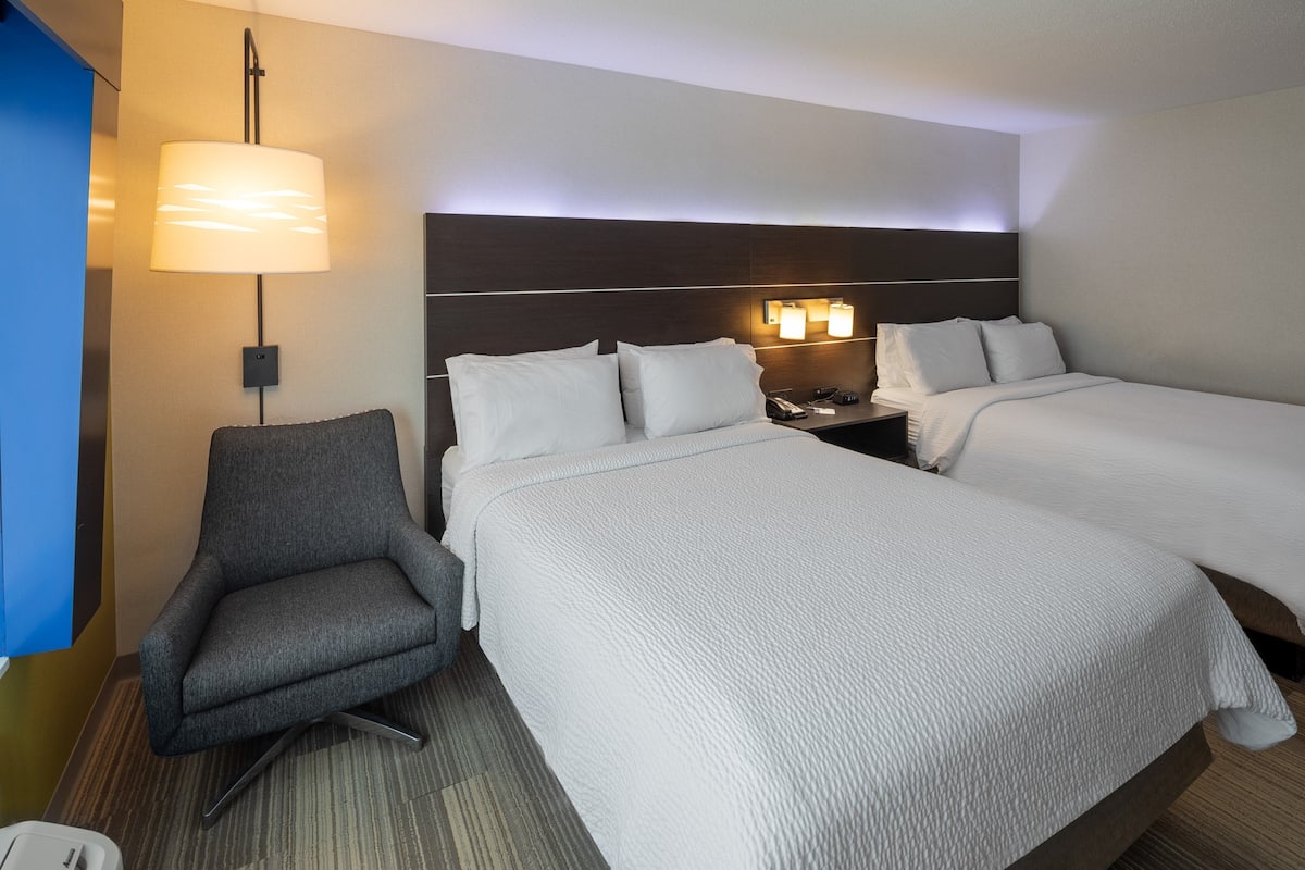 A hotel room with two beds, a desk, a chair, and a wall-mounted lamp.