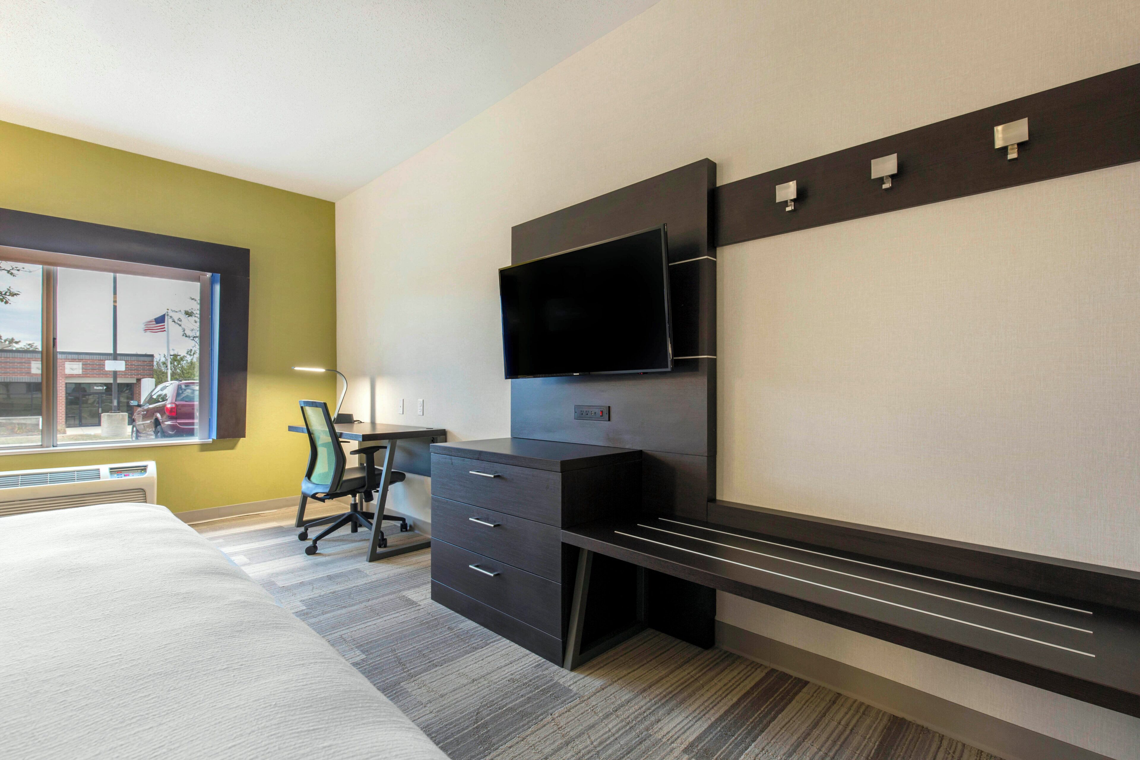 A hotel room with a bed, a desk with a chair, a flat-screen TV mounted on the wall, and a window with a view of the outside.