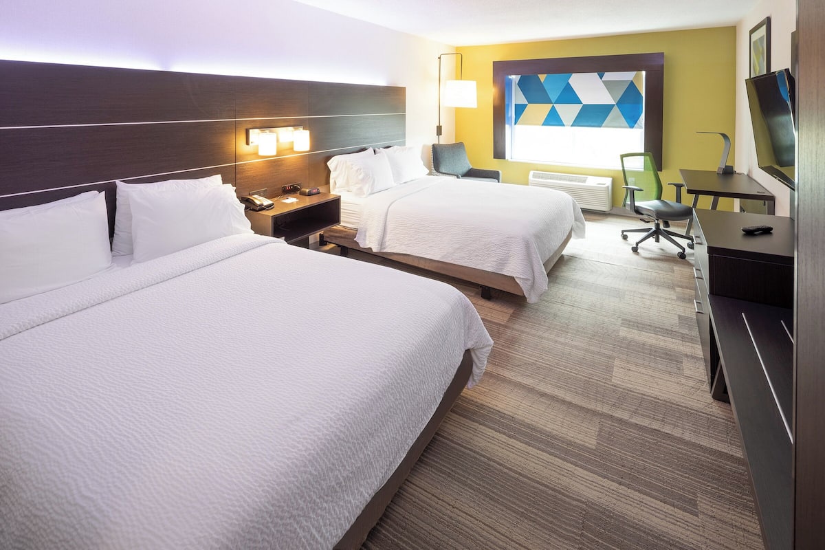 A hotel room with two beds, a desk, a chair, a TV, and a wall with geometric artwork.
