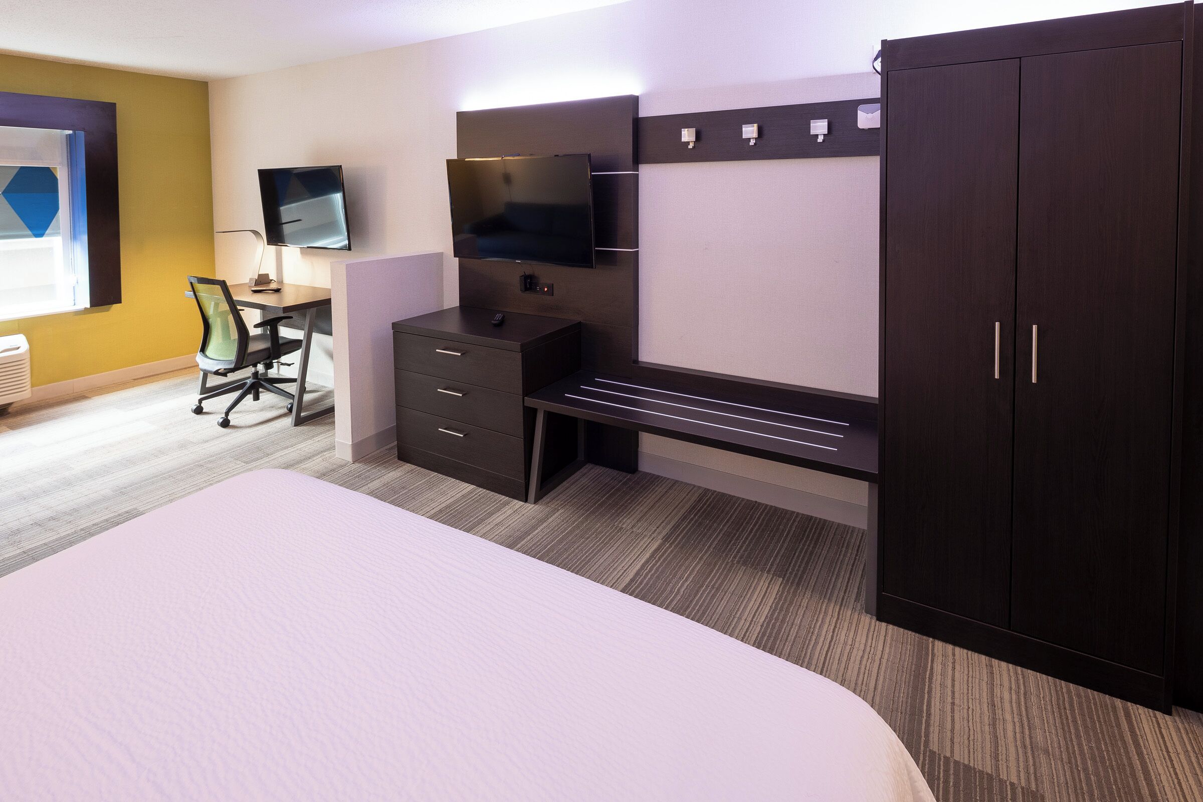 A hotel room with a bed, a desk with a chair, a TV mounted on the wall, and a wardrobe.