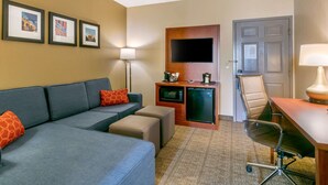Une chambre d’hôtel équipée d’un canapé, d’un pouf, d’un bureau, d’une chaise, d’un téléviseur à écran plat, d’un four à micro-ondes, d’un réfrigérateur, d’une lampe et de tableaux encadrés accrochés au mur.