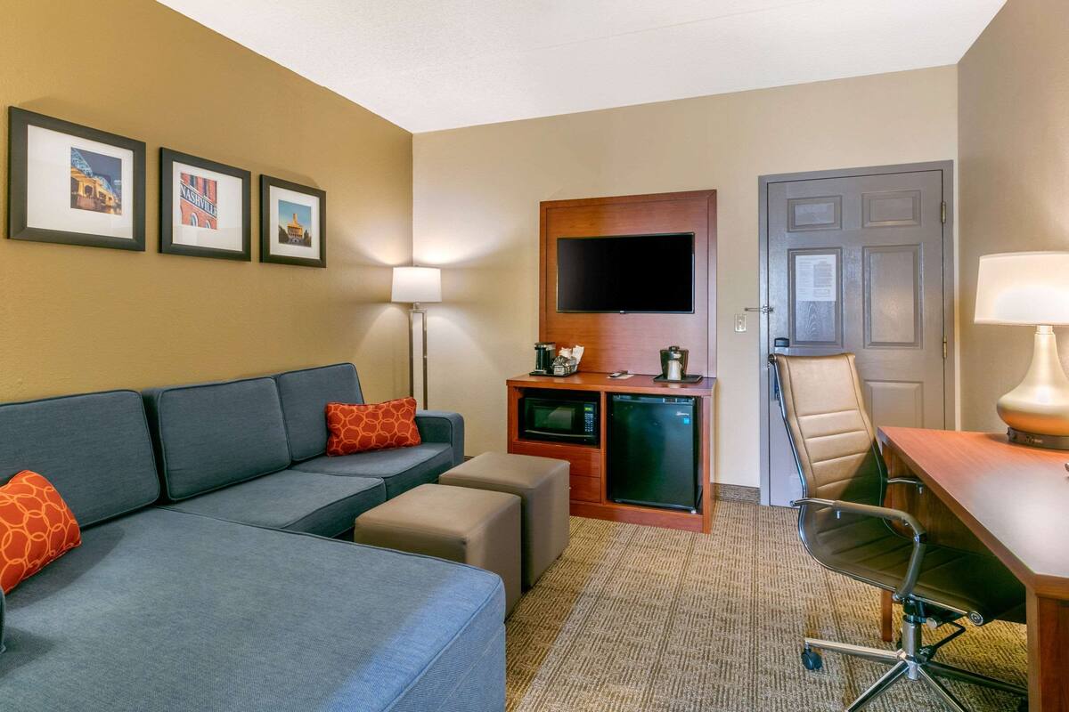 Habitación de hotel con sofá, reposapiés, escritorio, silla, televisor de pantalla plana, microondas, refrigerador, lámpara y cuadros en la pared.