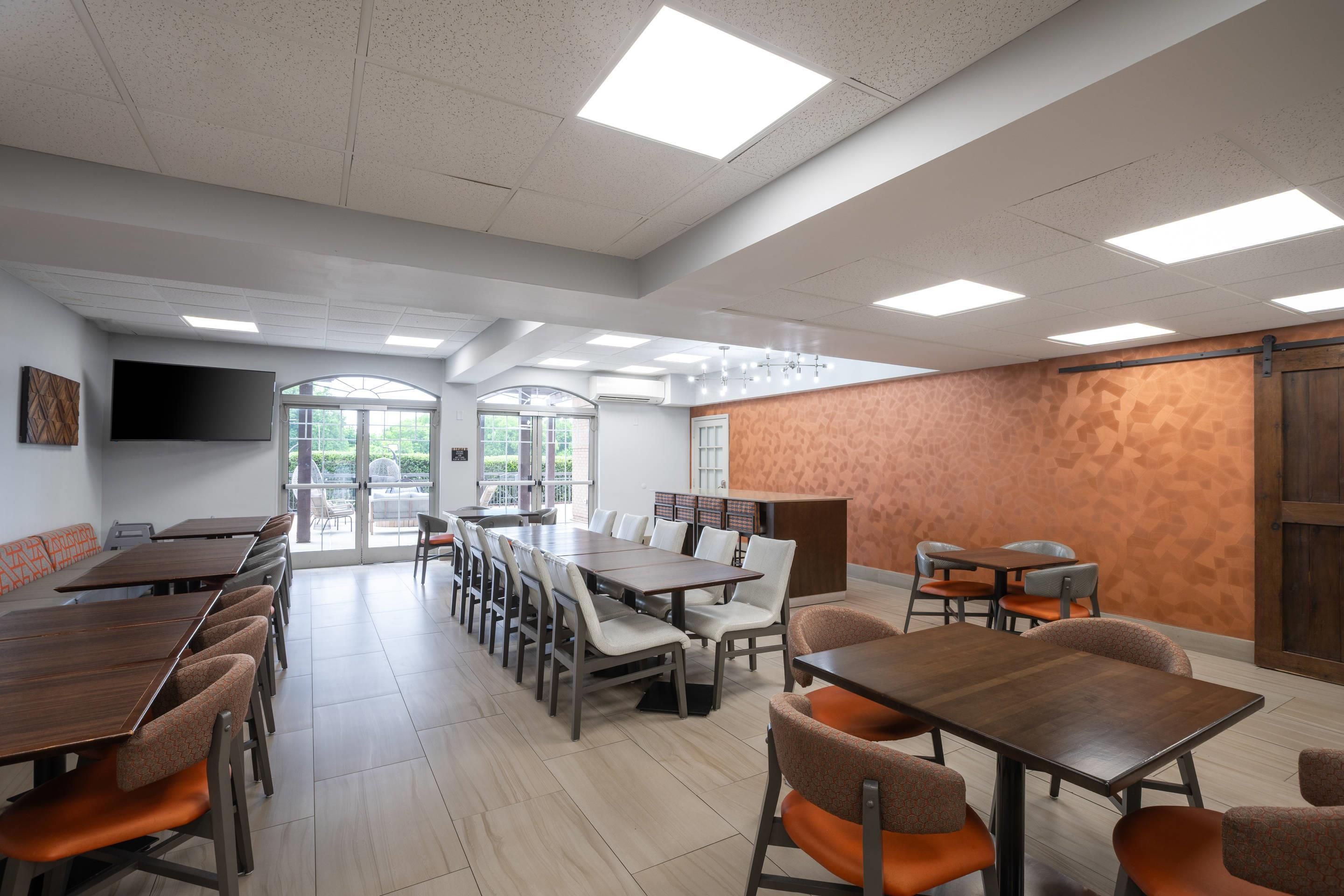 A spacious dining area with multiple tables and chairs, large windows, and a flat-screen TV mounted on the wall.