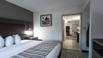 Una habitación de hotel con una cama grande, una mesita de noche con lámpara, una obra de arte abstracta enmarcada en la pared y vistas a la habitación contigua.