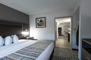 A hotel room with a large bed, a bedside table with a lamp, a framed abstract artwork on the wall, and a view into the adjacent room.