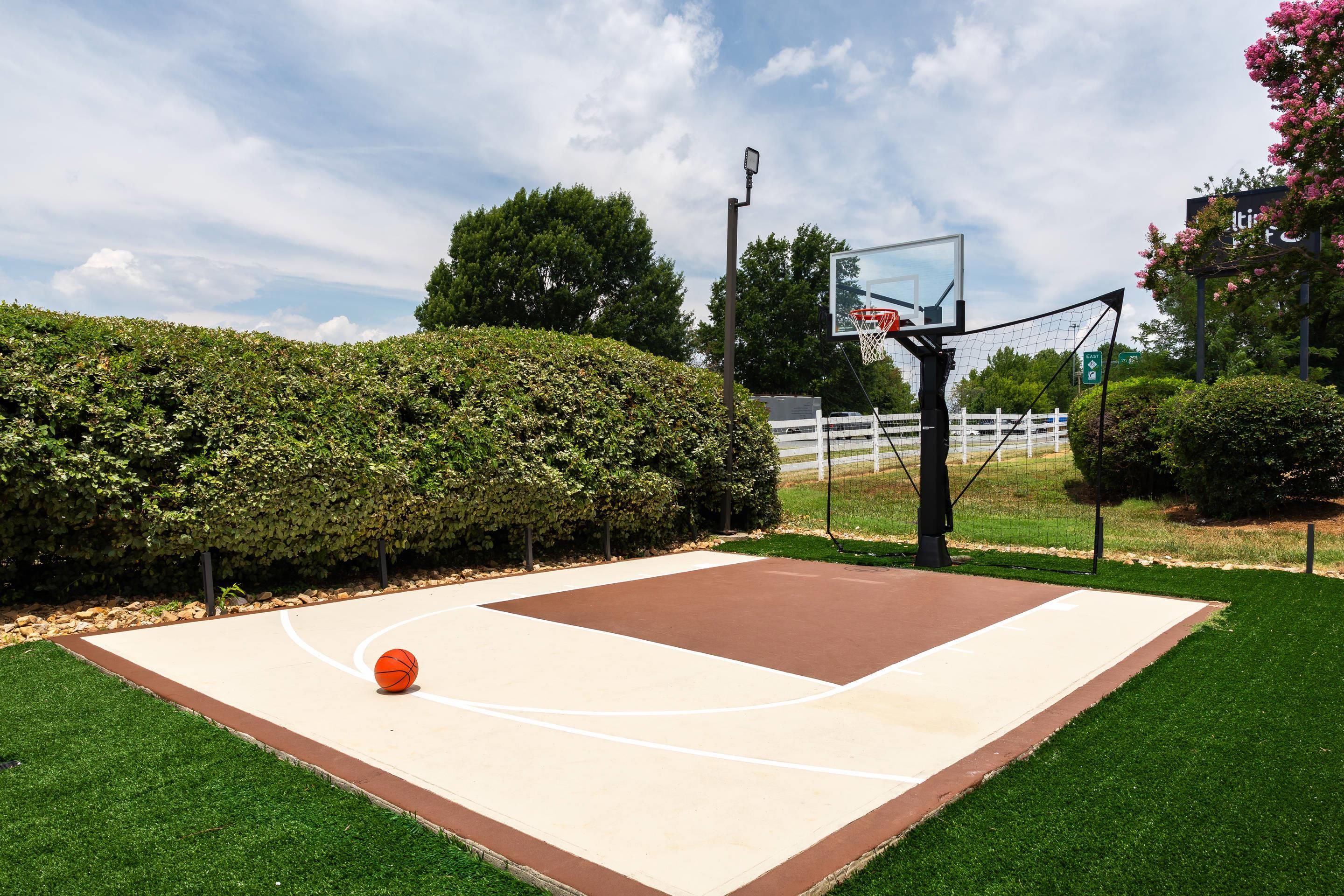 En utomhusbasketplan med en basketkorg, omgiven av välskött gräs och häckar.