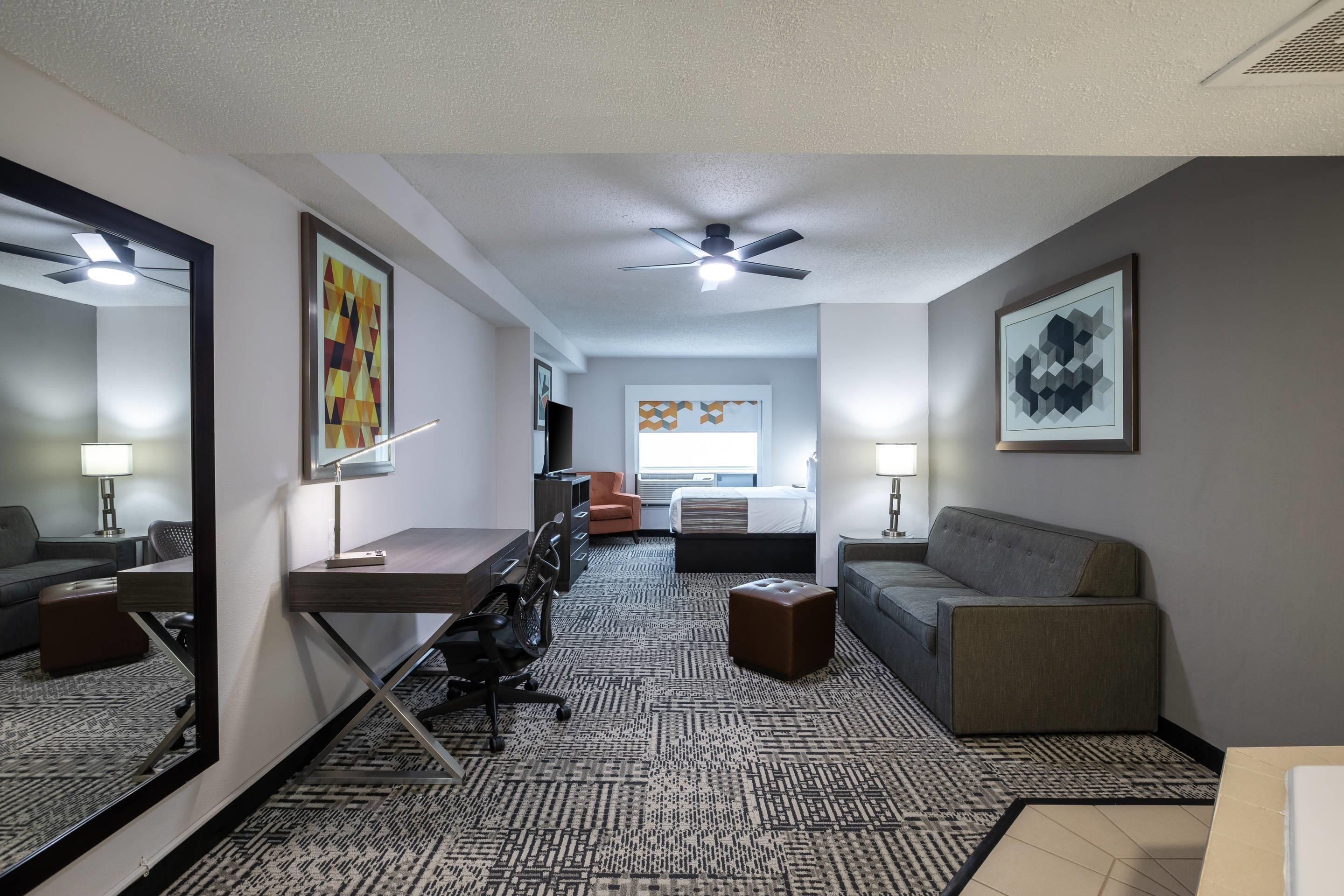 A hotel room with a bed, a desk, a chair, a sofa, a mirror, and a ceiling fan.
