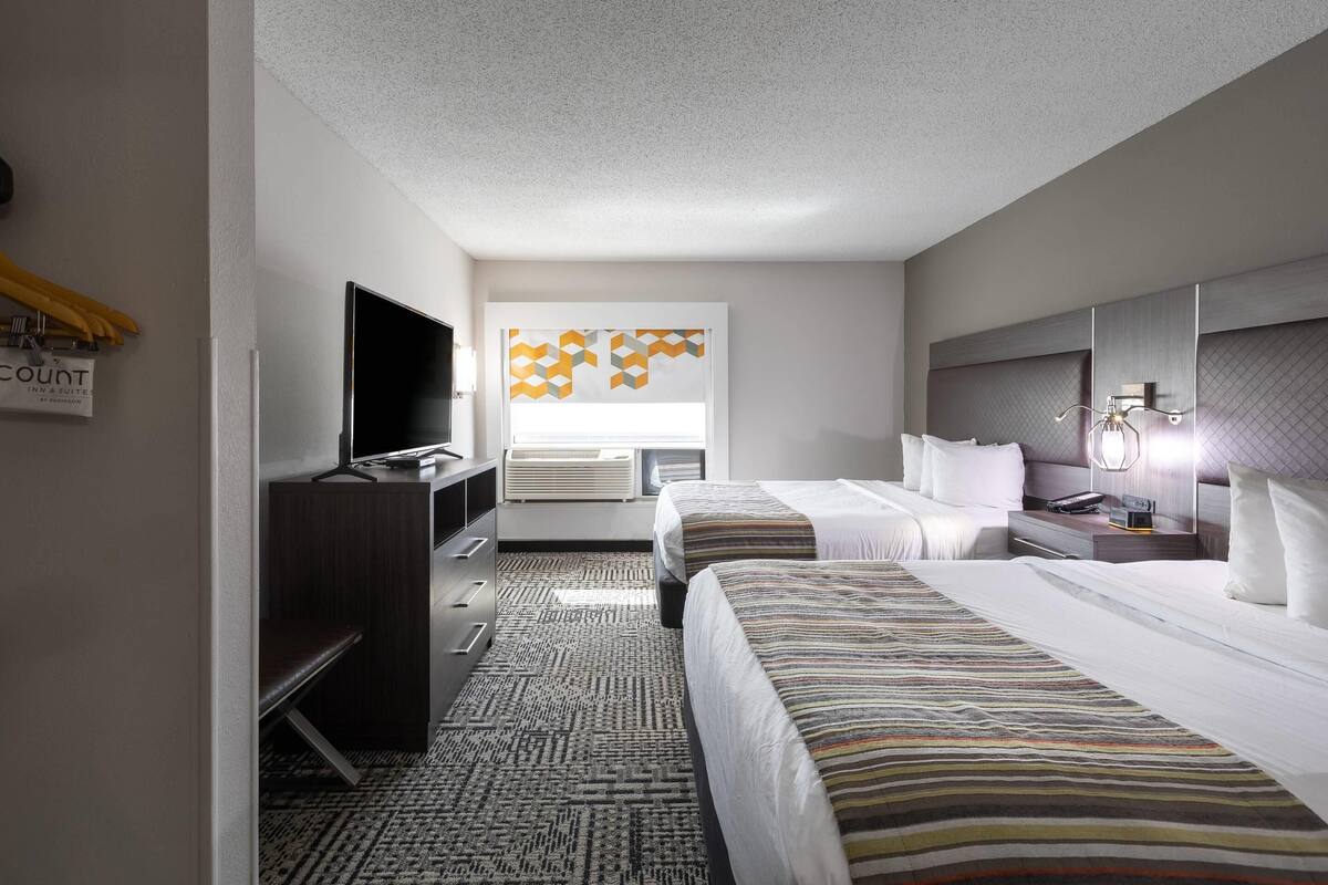 A hotel room with two beds, a TV, a desk, and a window with geometric decor.