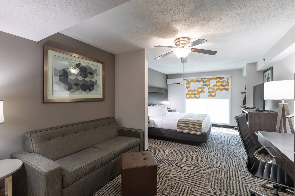 A hotel room with a sofa, a small ottoman, a desk with a chair, a bed with a striped bedspread, a ceiling fan, and a geometric-patterned wall hanging.