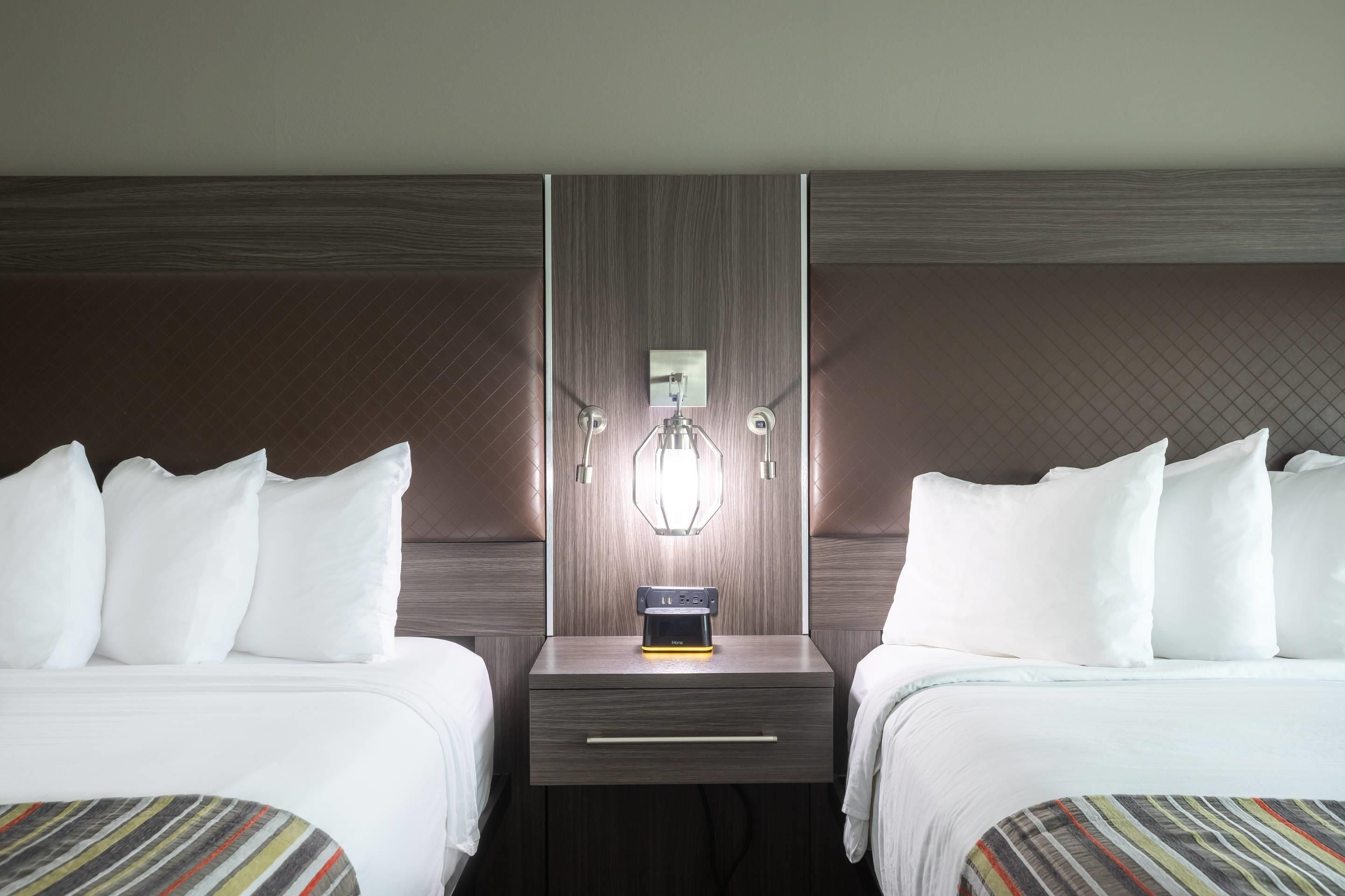 A hotel room with two beds, a nightstand with a lamp, and a wall-mounted control panel.
