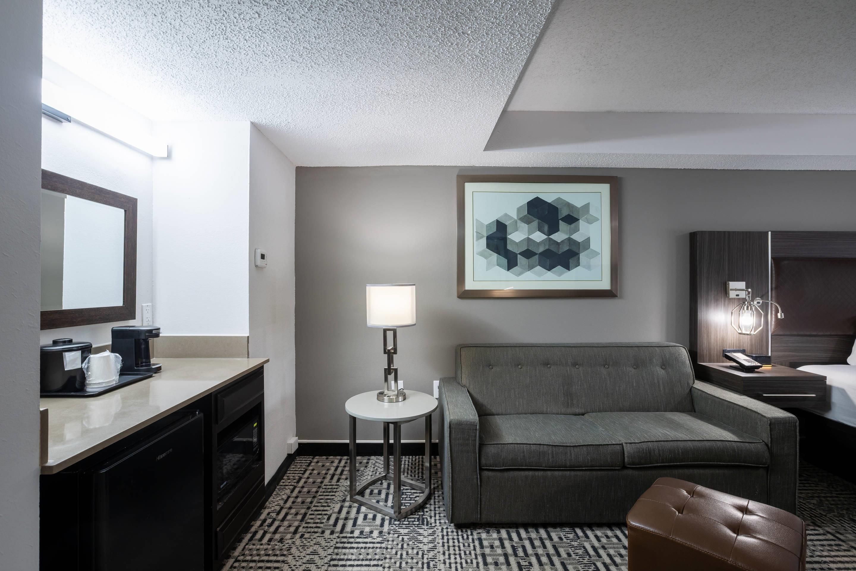 Una habitación de hotel con sofá, una mesita auxiliar, lámpara, cama, mesita de noche, espejo y un cuadro en la pared.