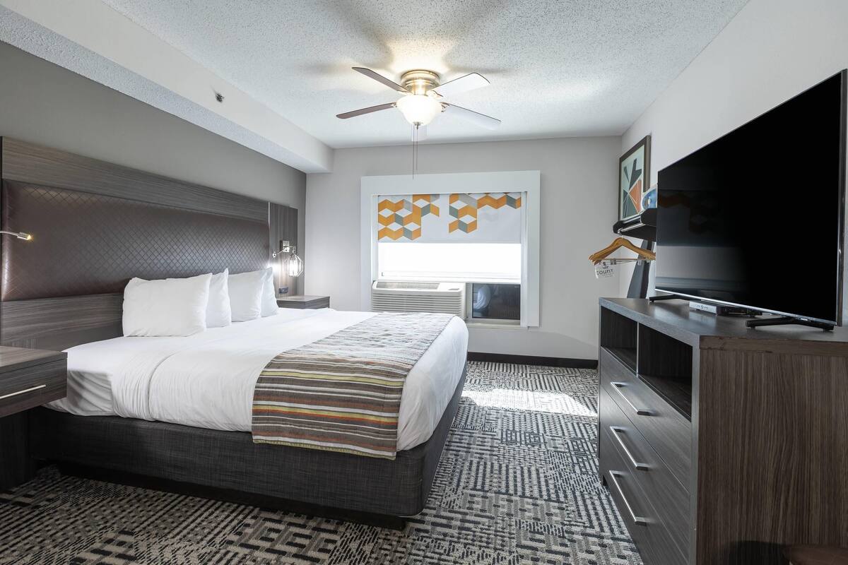 A hotel room with a bed, a television, a ceiling fan, a window with geometric curtains, and a patterned rug.