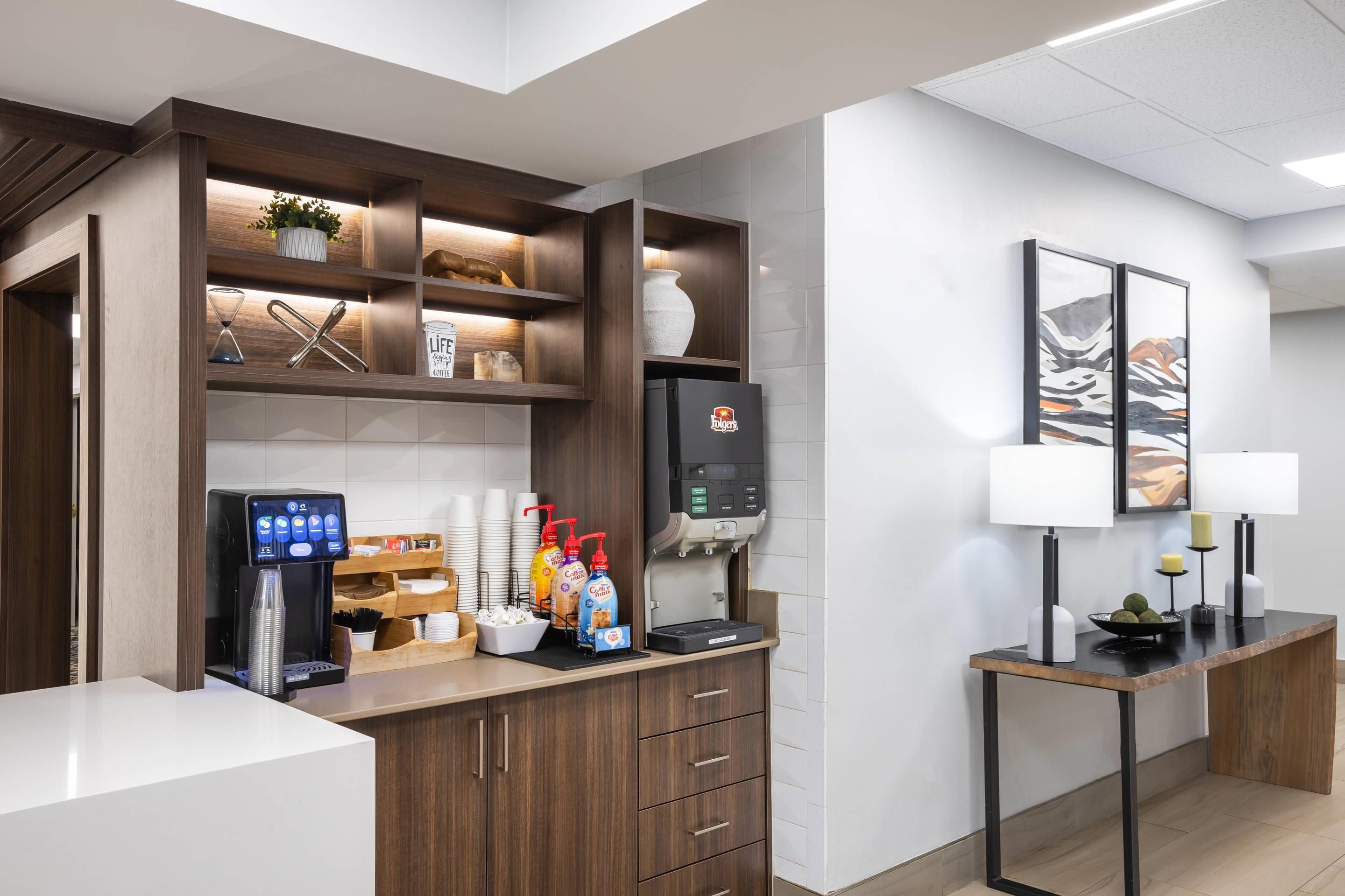Un área de recepción de hotel moderna con una máquina de café, dispensadores de bebidas y una pantalla digital.