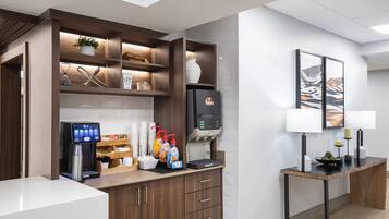 Un área de recepción de hotel moderna con una máquina de café, dispensadores de bebidas y una pantalla digital.